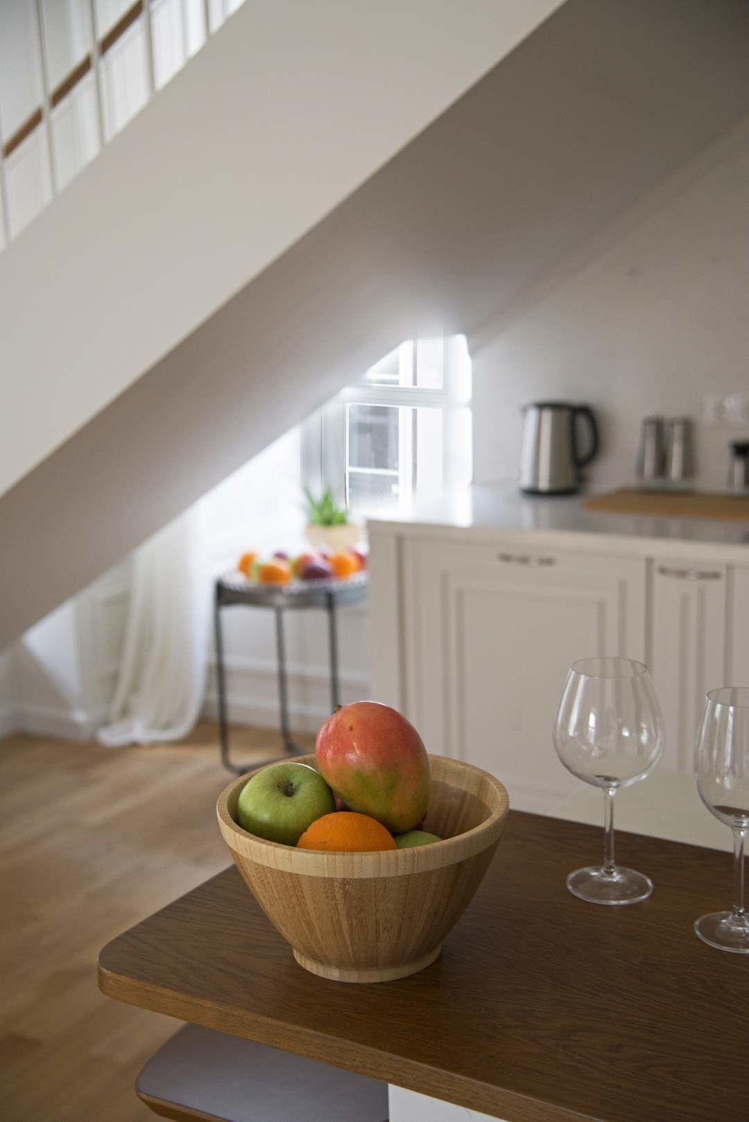 A wooden bowl with fruits on a table in a kitchen with white cabinets and a window.