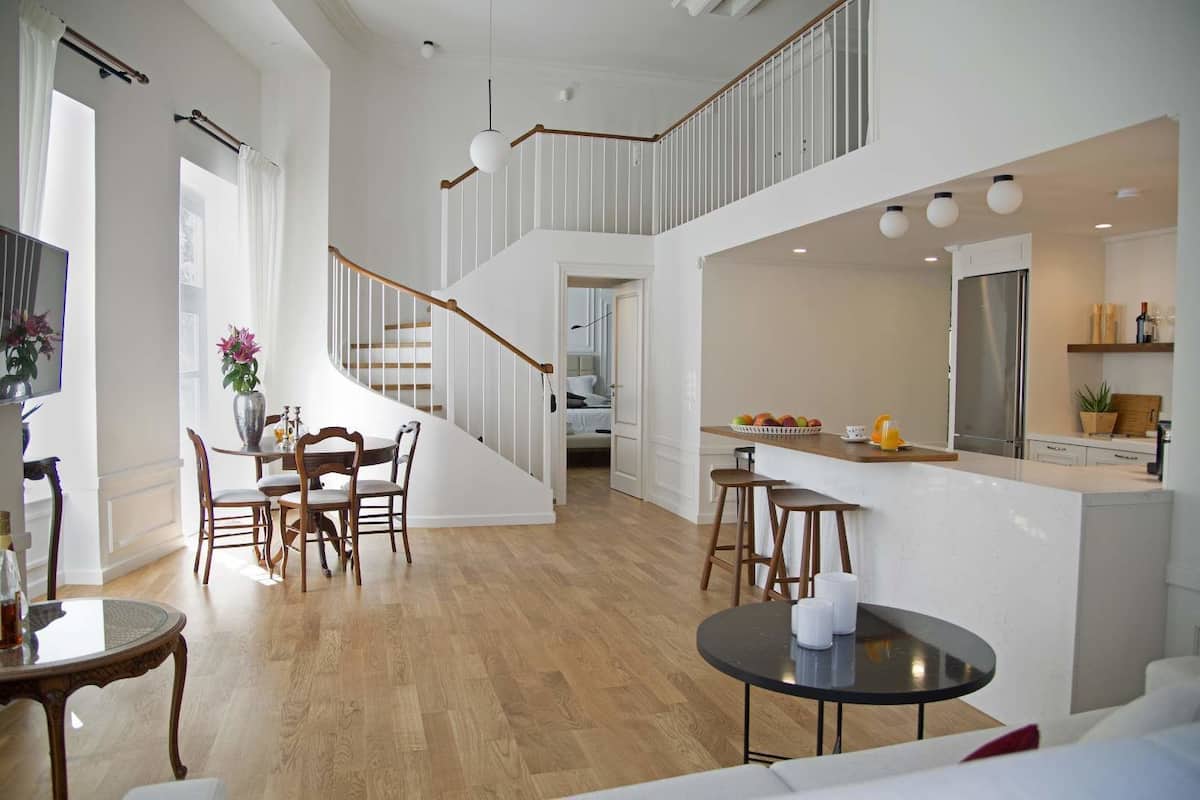 A spacious living area with a wooden floor, a dining table with chairs, a kitchen area with a counter and stools, and a staircase leading to an upper level.