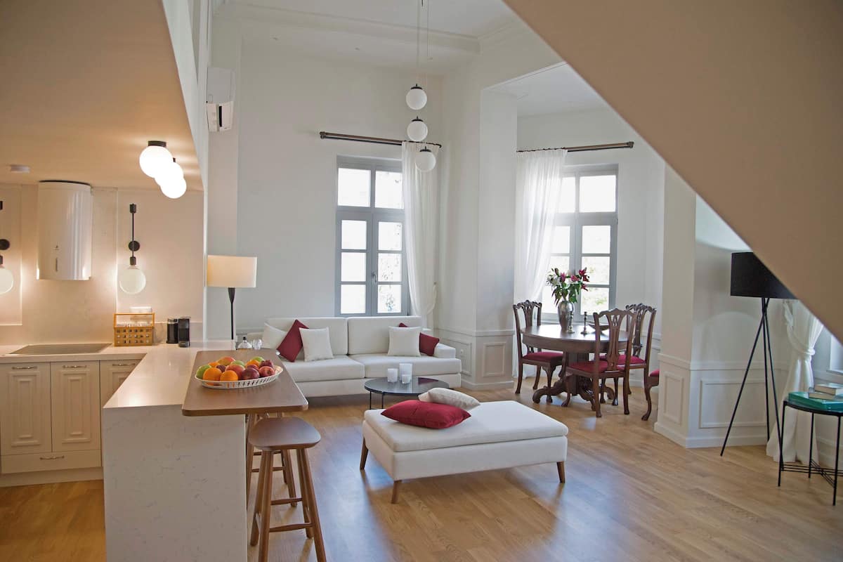 A spacious living area with a white sofa, ottoman, and a dining table with chairs. A kitchen area is visible on the left with a fruit bowl on the counter.