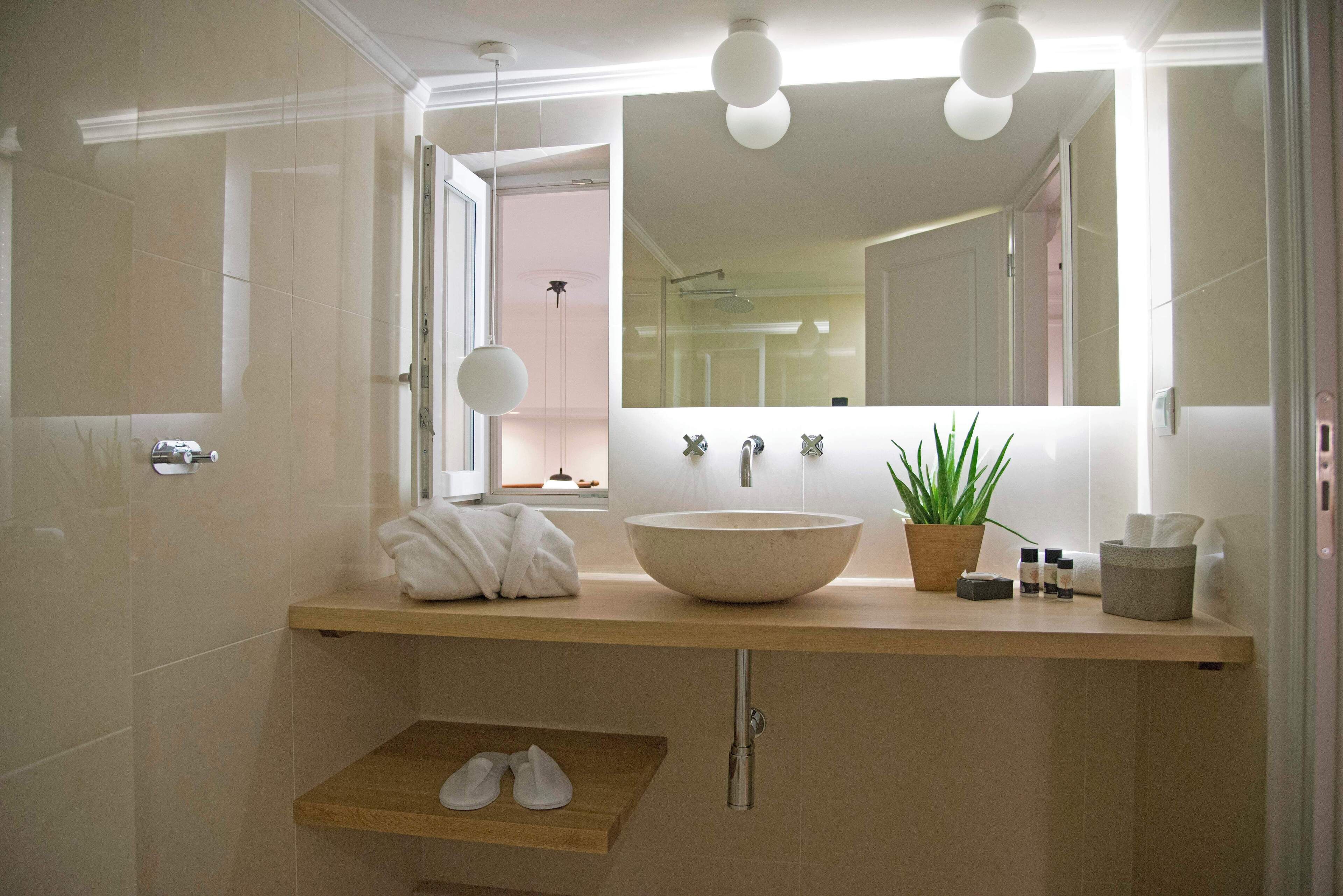 Une salle de bain moderne avec un grand miroir, un lavabo sur colonne, une plante en pot et une étagère en bois où sont posées des chaussures.