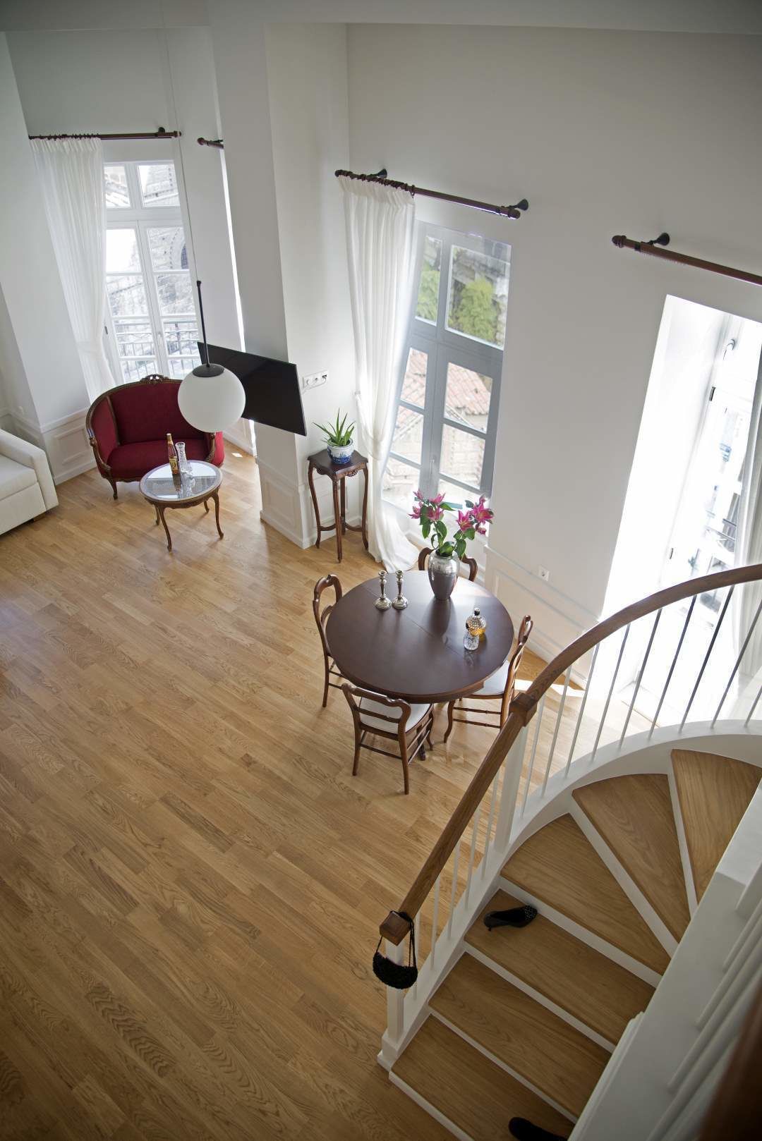 A spacious living room with a wooden floor, a red sofa, a round dining table with chairs, a staircase, and large windows with curtains.