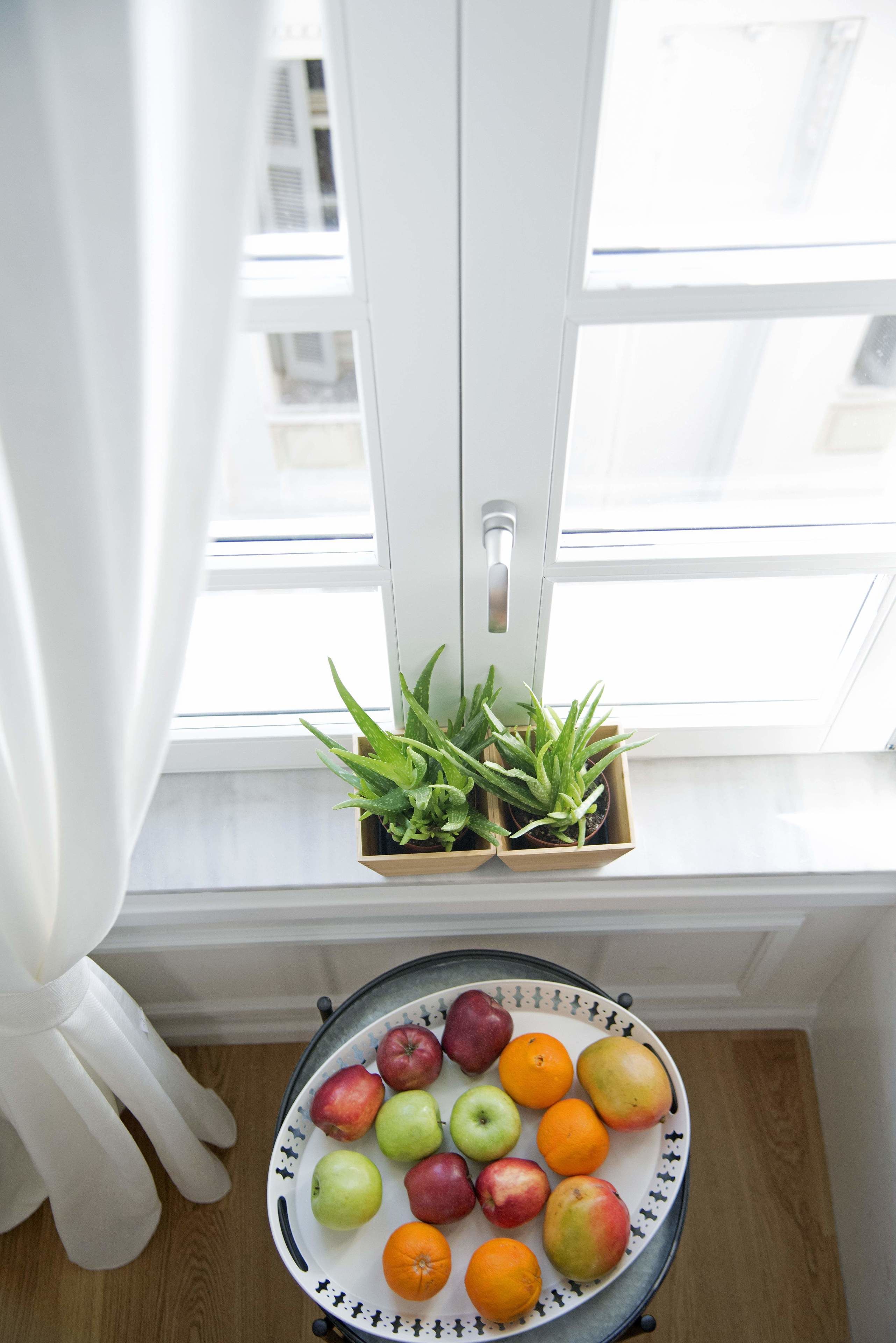 A window with a potted plant, a fruit bowl with apples and oranges, and a white chair.