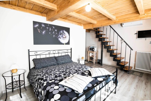 A bedroom with a bed, a black and white polka dot bedspread, a black metal headboard, a small round table with a lamp, a black and white wall art, a staircase, and a white radiator. - Agriturismo Cà del Nono (Moasca)