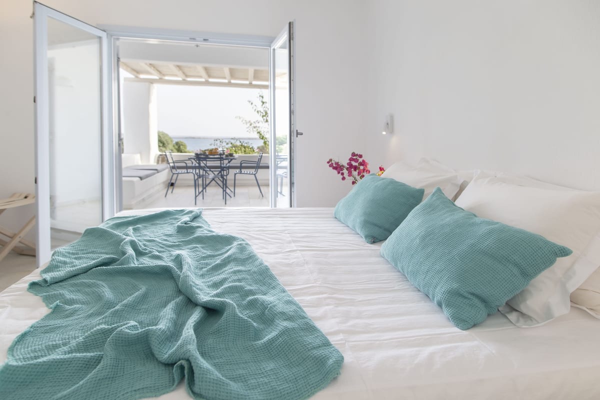 A bedroom with a bed, a teal blanket, and pillows. There is a view of an outdoor area with a table and chairs.