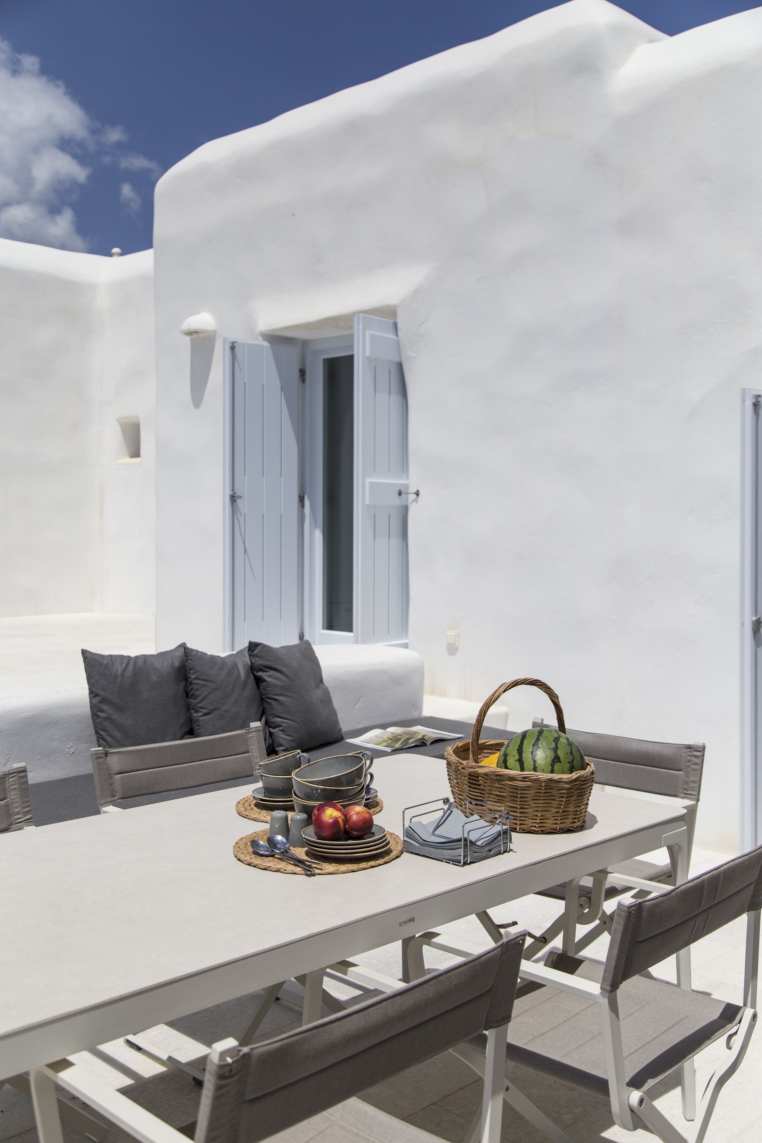 Une pièce aux murs blancs, avec une table dressée pour le repas, agrémentée d’un panier de fruits et d’une pastèque.