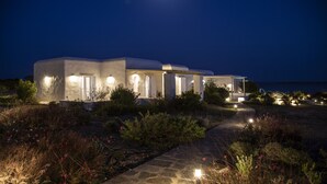 A modern, white building with multiple rooms, a stone pathway, and a view of the sea at night. - The Sand Collection (Paros)
