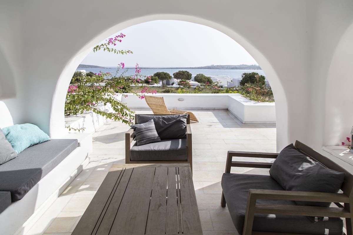 A sunlit outdoor seating area with a view of the sea, featuring a wooden table and two sofas with cushions.