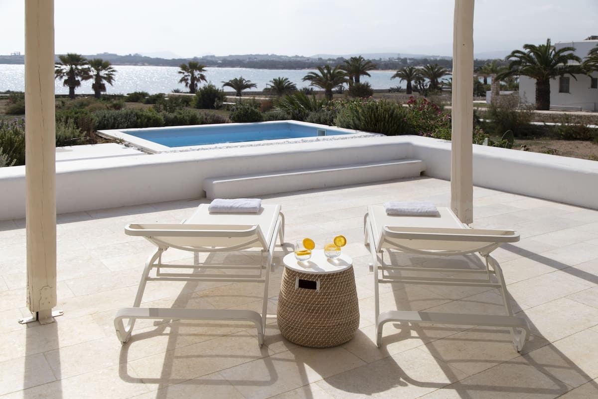 A sunlit outdoor area with a pool, two lounge chairs, and a small table with a basket.