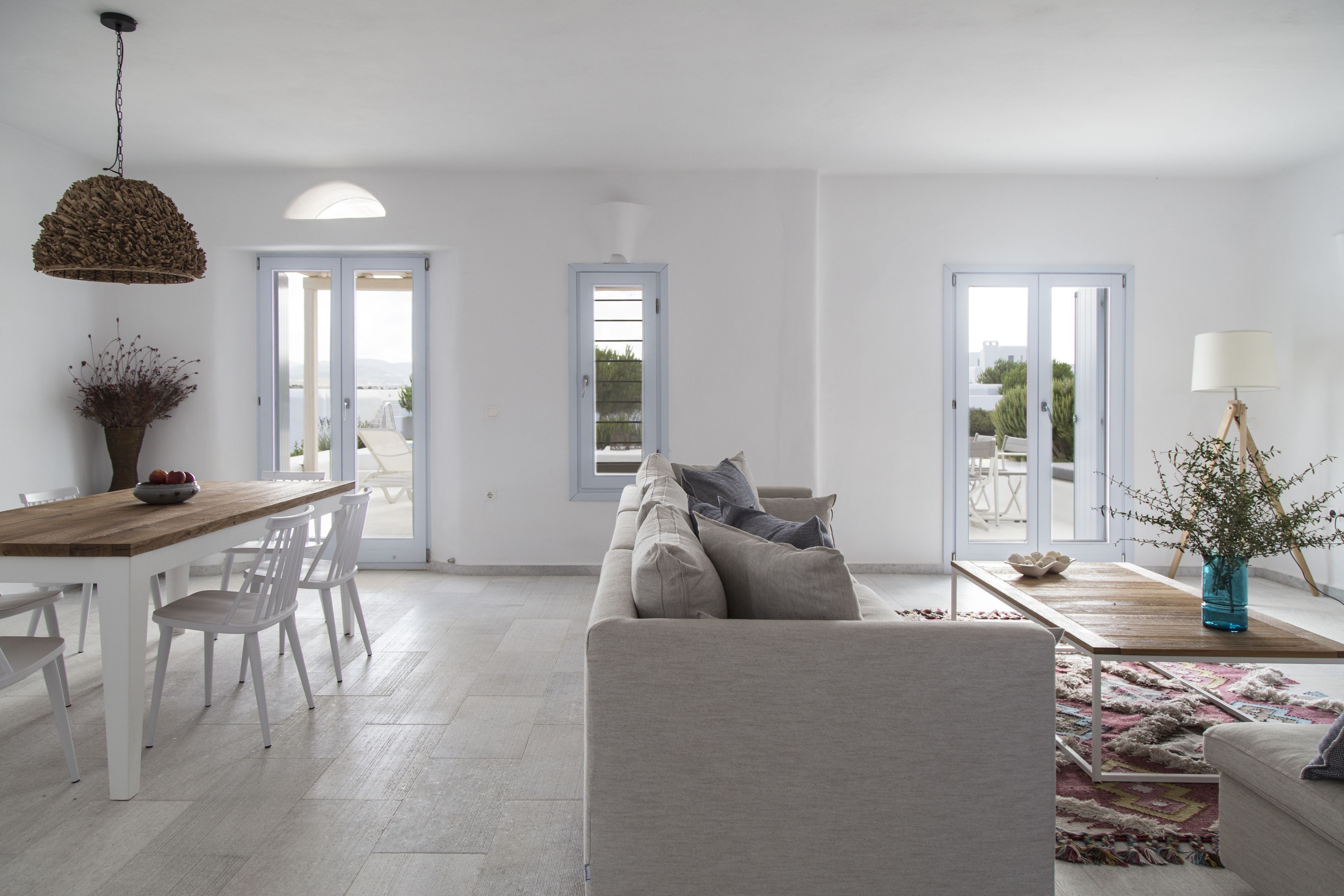 Een moderne woonkamer met een houten eettafel, witte stoelen, een beige bank en een salontafel. Er zijn twee deuren die leiden naar buitenruimtes.