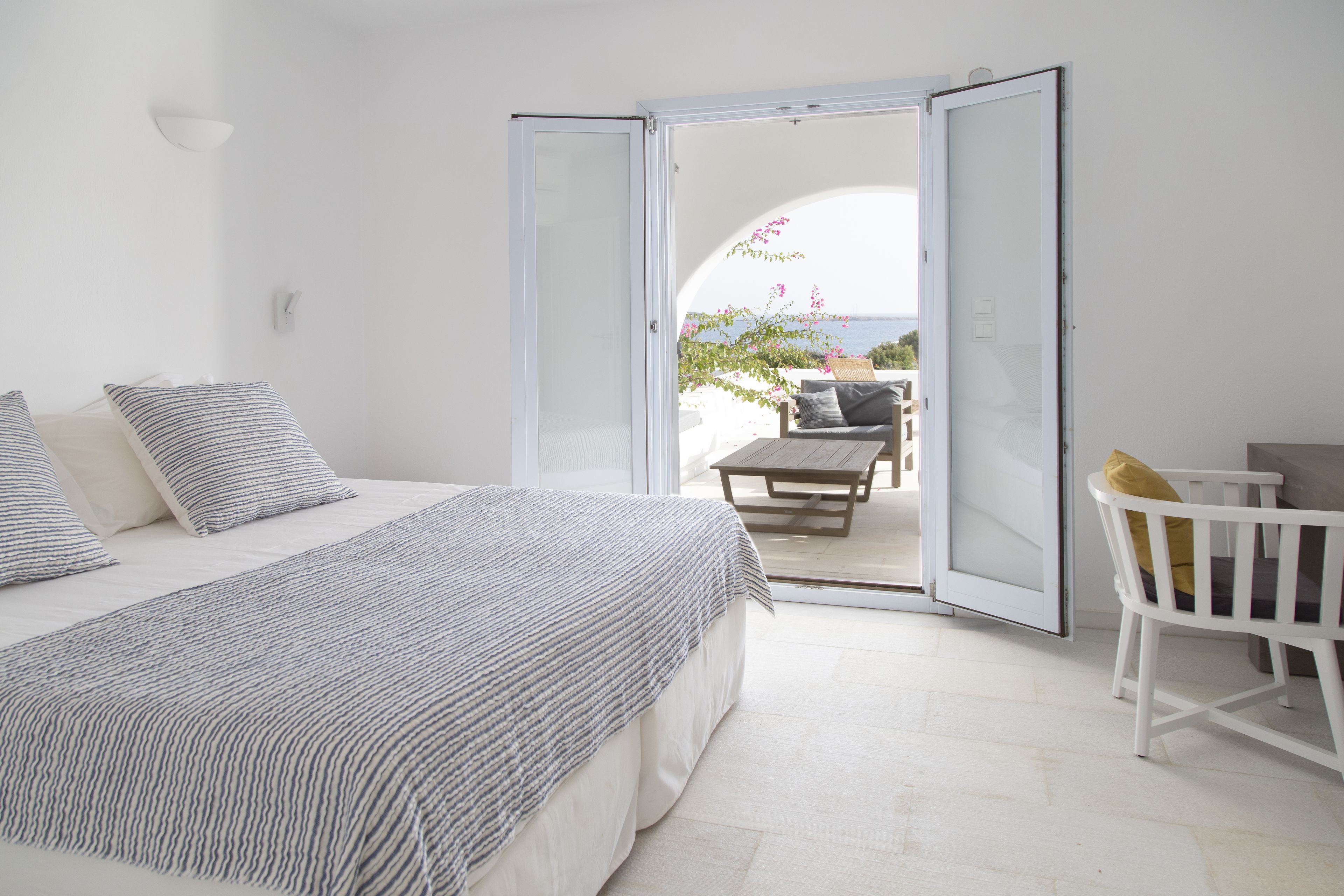 A bedroom with a bed, a bedside table, and a chair. There is a sliding glass door leading to an outdoor area with a table and chairs.
