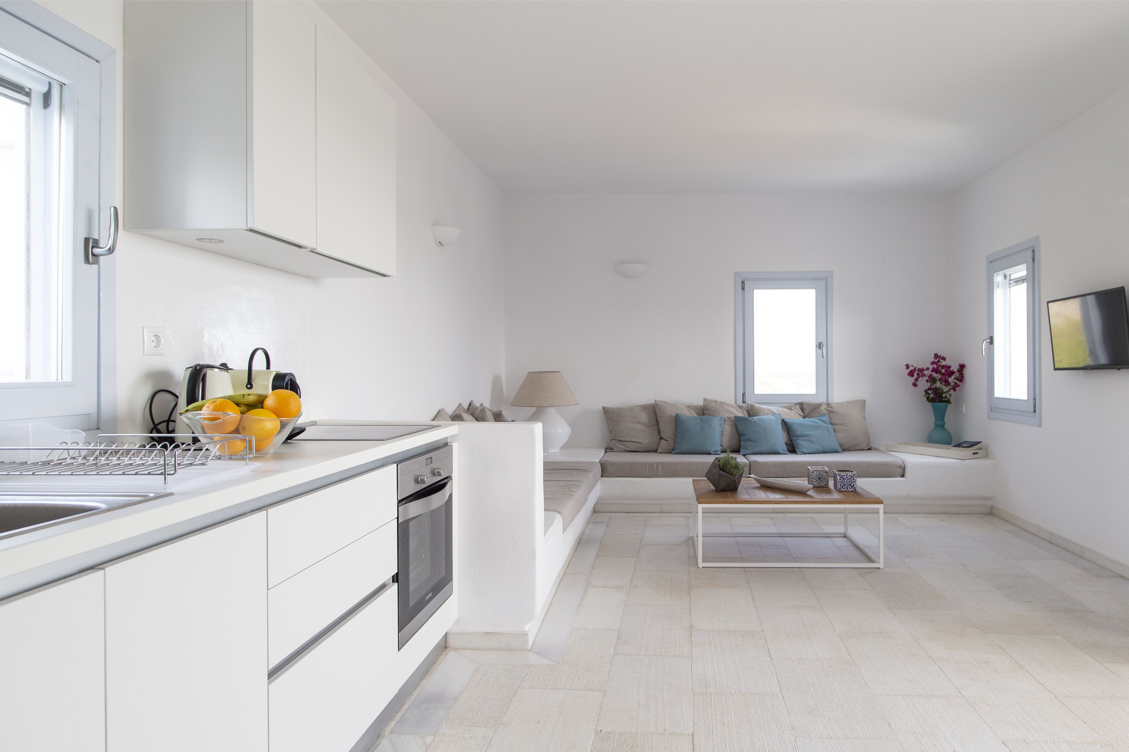 A modern kitchen with white cabinetry, a built-in oven, and a sink. There is a dining area with a white table and a sofa with cushions. A television is mounted on the wall, and a window provides natural light.