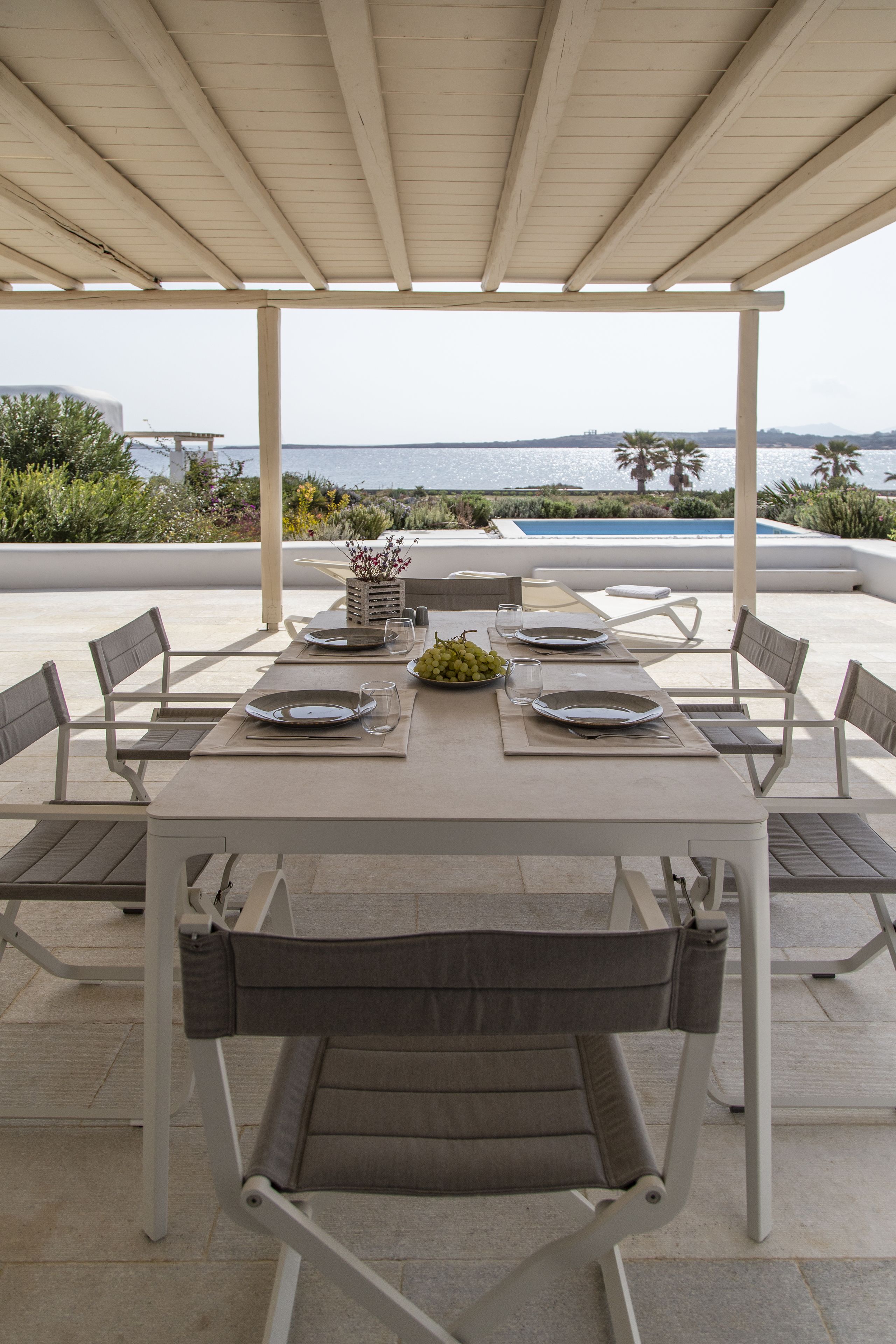 Un área de comedor al aire libre cubierta, con una mesa preparada para cuatro, con vistas a una piscina y un paisaje pintoresco.