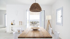 A dining area with a wooden table, white chairs, a hanging light fixture, and a view through an open door. - The Sand Collection (Paros)