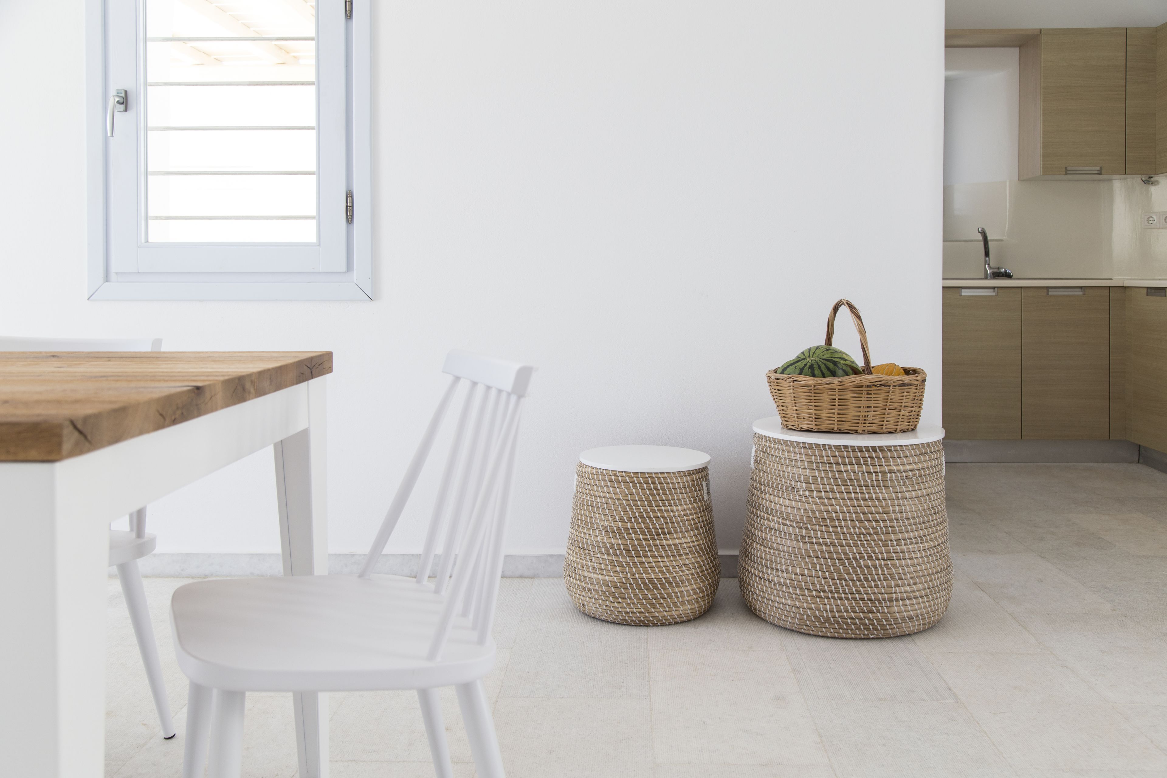 Una cocina moderna con armarios blancos, encimera de madera y una silla blanca. Hay dos taburetes trenzados y una cesta con frutas sobre la encimera.