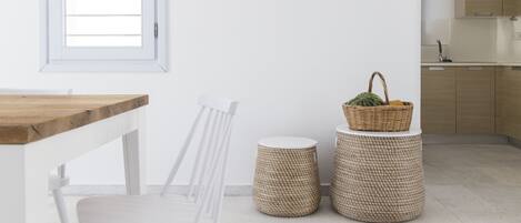 Una cocina moderna con armarios blancos, encimera de madera y una silla blanca. Hay dos taburetes trenzados y una cesta con frutas sobre la encimera.