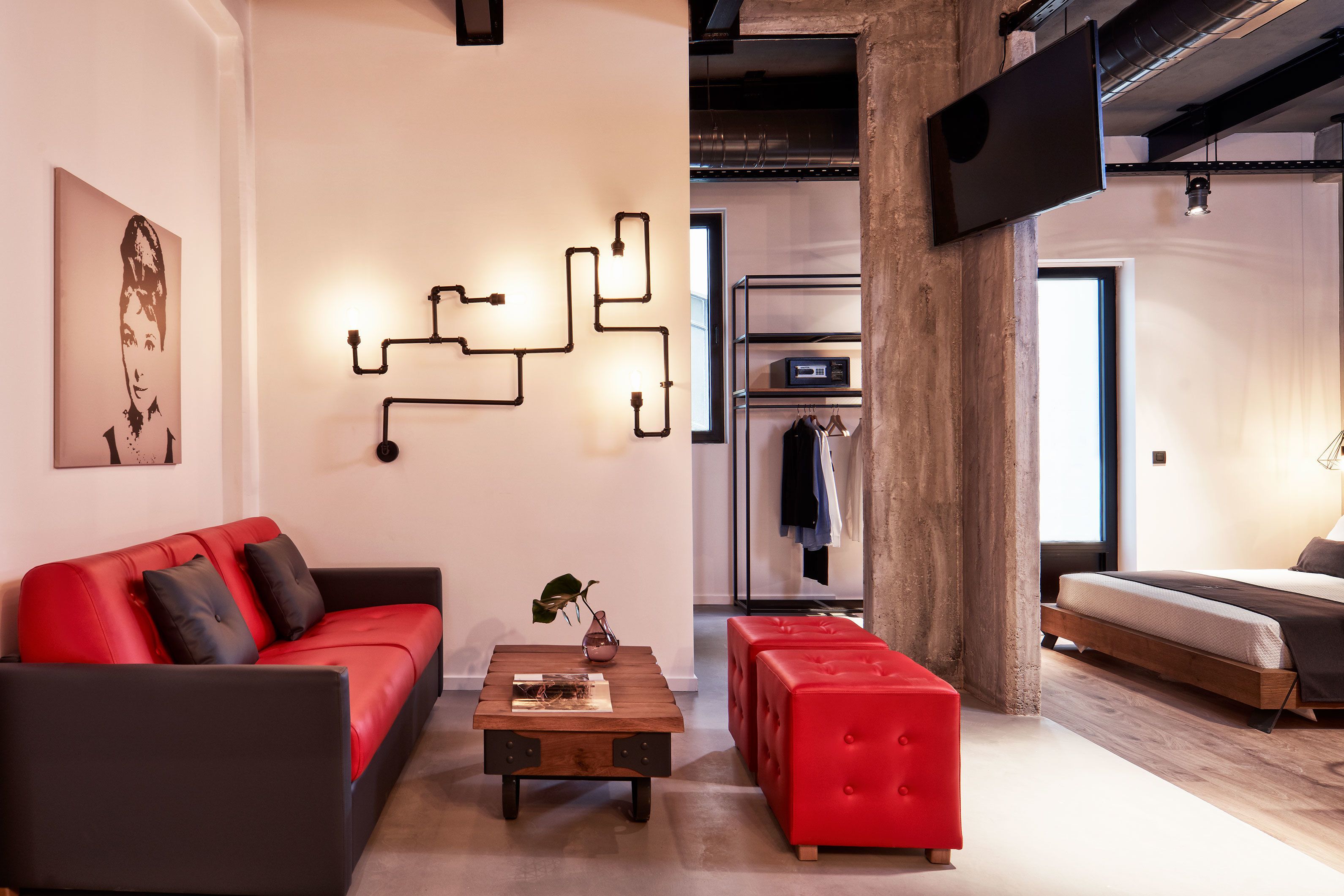 Uma sala de estar moderna com um sofá vermelho, uma mesa de centro de madeira e um banquinho vermelho. Há um luminário fixado na parede e uma grande tela de televisão.