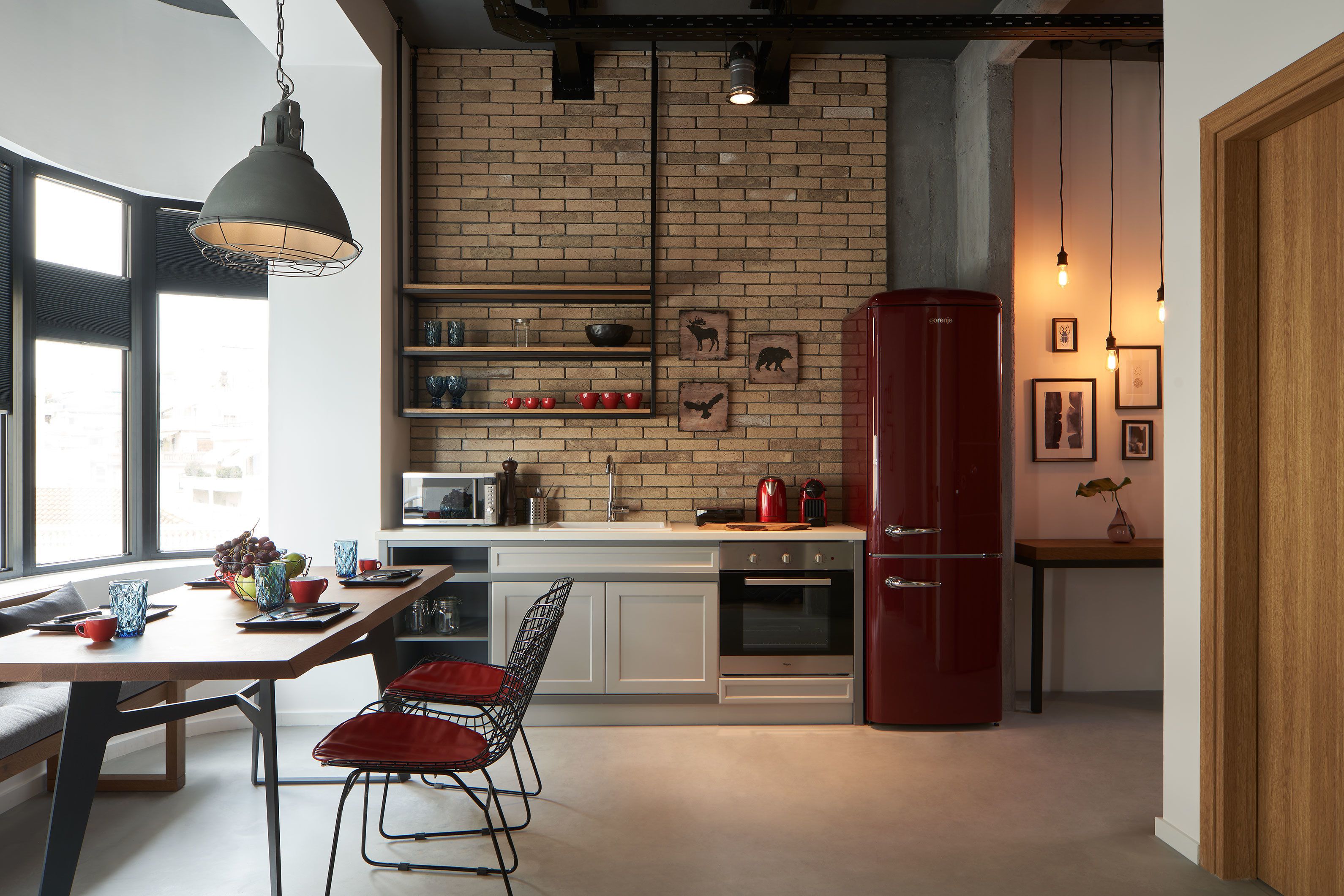 A modern kitchen with a brick wall, white cabinets, a red refrigerator, and a dining area with red chairs.