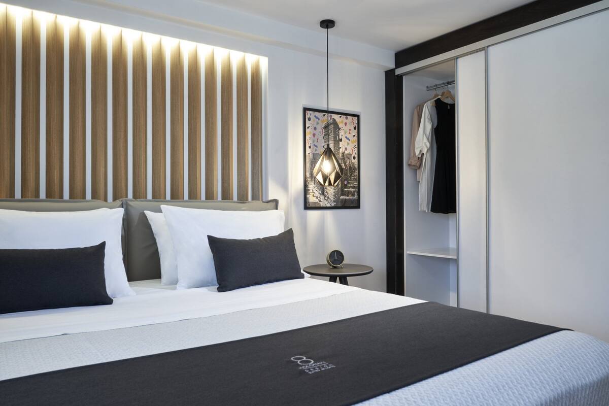 Ein modernes Hotelzimmer mit einem großen Bett, einer Kopfteil mit vertikalen Lamellen, einem gerahmten Stadtbild und einer hängenden Leuchte.