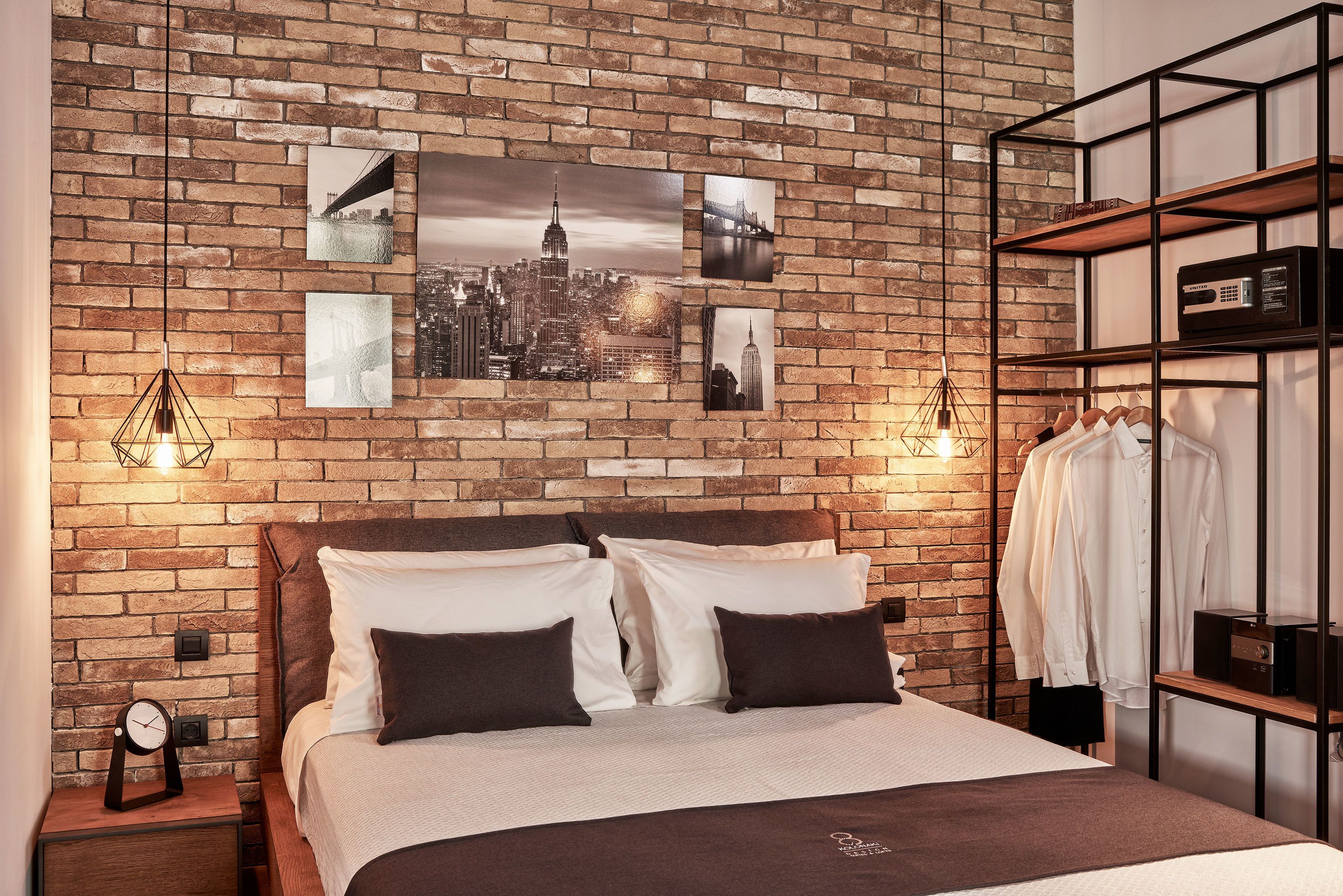 Ein Schlafzimmer mit einer Ziegelwand, einem Bett mit weißer und brauner Bettwäsche, einem Nachttisch mit einer Uhr und einem Wandregal mit Kleidung.