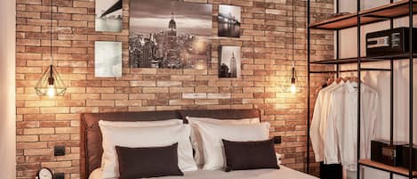 Ein Schlafzimmer mit einer Ziegelwand, einem Bett mit weißer und brauner Bettwäsche, einem Nachttisch mit einer Uhr und einem Wandregal mit Kleidung.