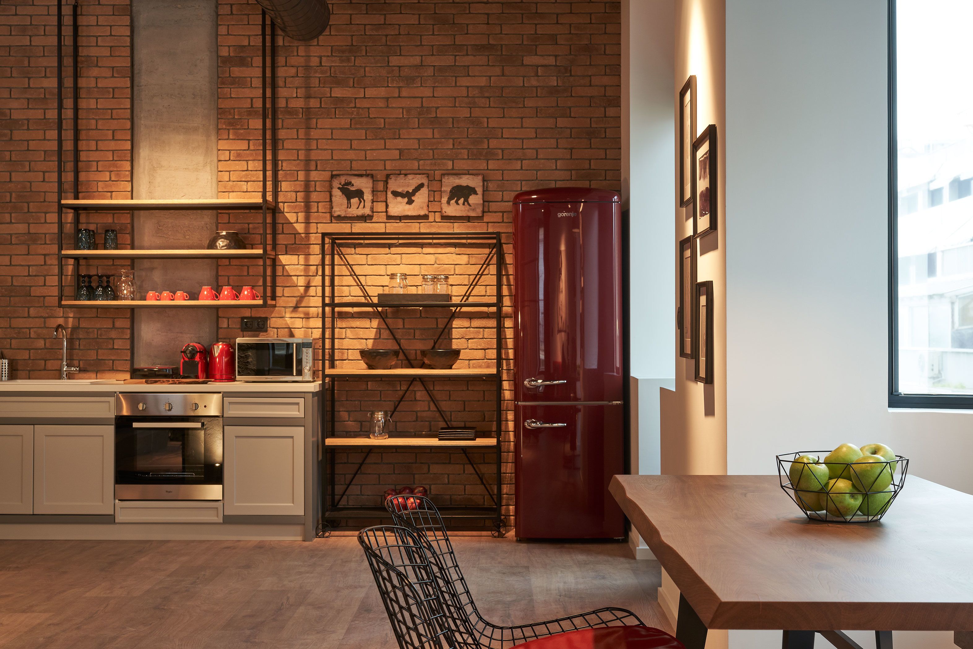 A kitchen with a brick wall, a red refrigerator, and a table with a bowl of apples.