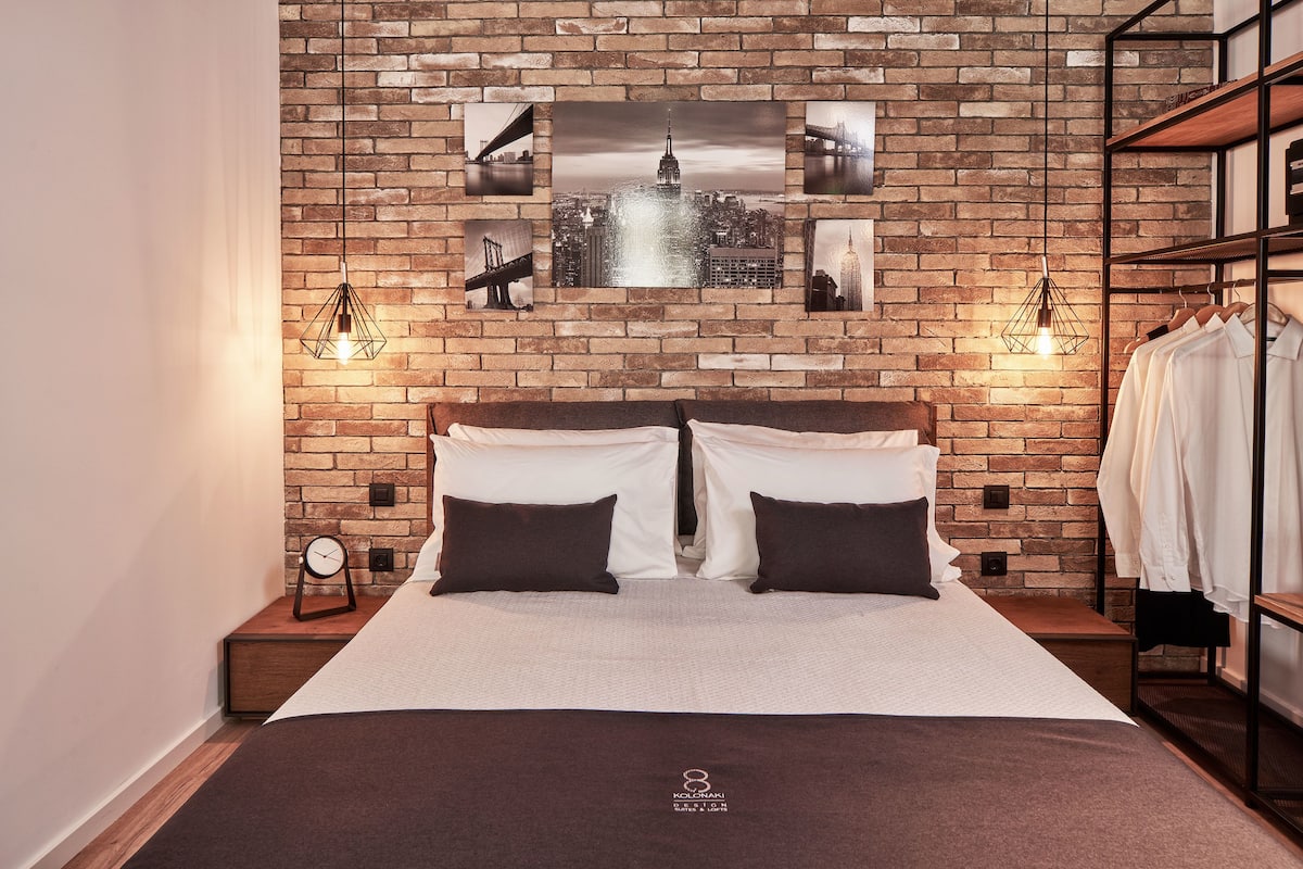 Ein Hotelzimmer mit Backsteinwand, einem Bett mit weißen und dunklen Kissen, zwei Wandlampen und einem Kleiderhaken mit aufgehängter Kleidung.