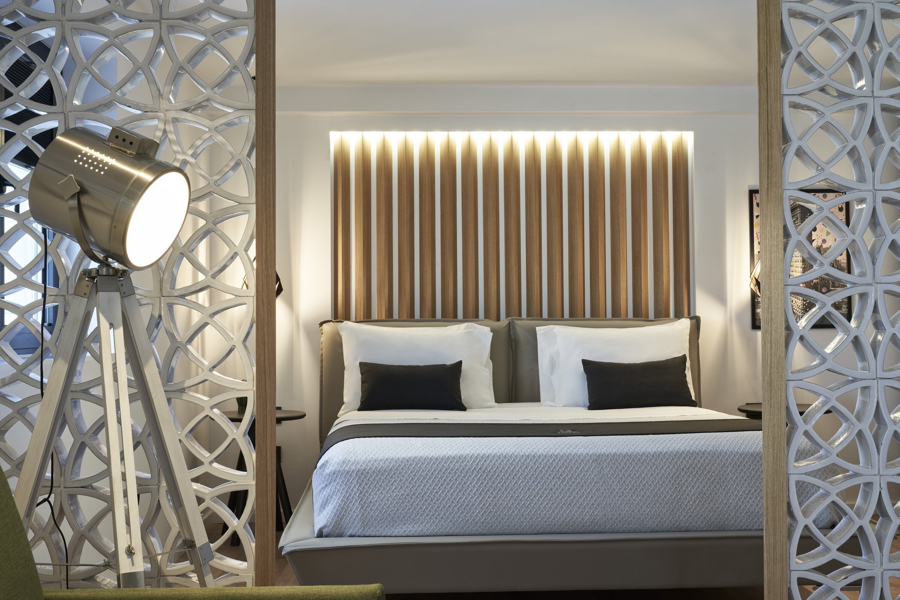 Quarto moderno com cama, mesa de cabeceira, luminária e painéis decorativos.