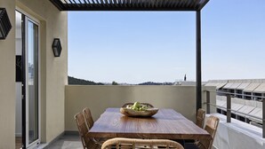 A balcony with a wooden table and wicker chairs, a bowl of fruit, and a view of the cityscape.