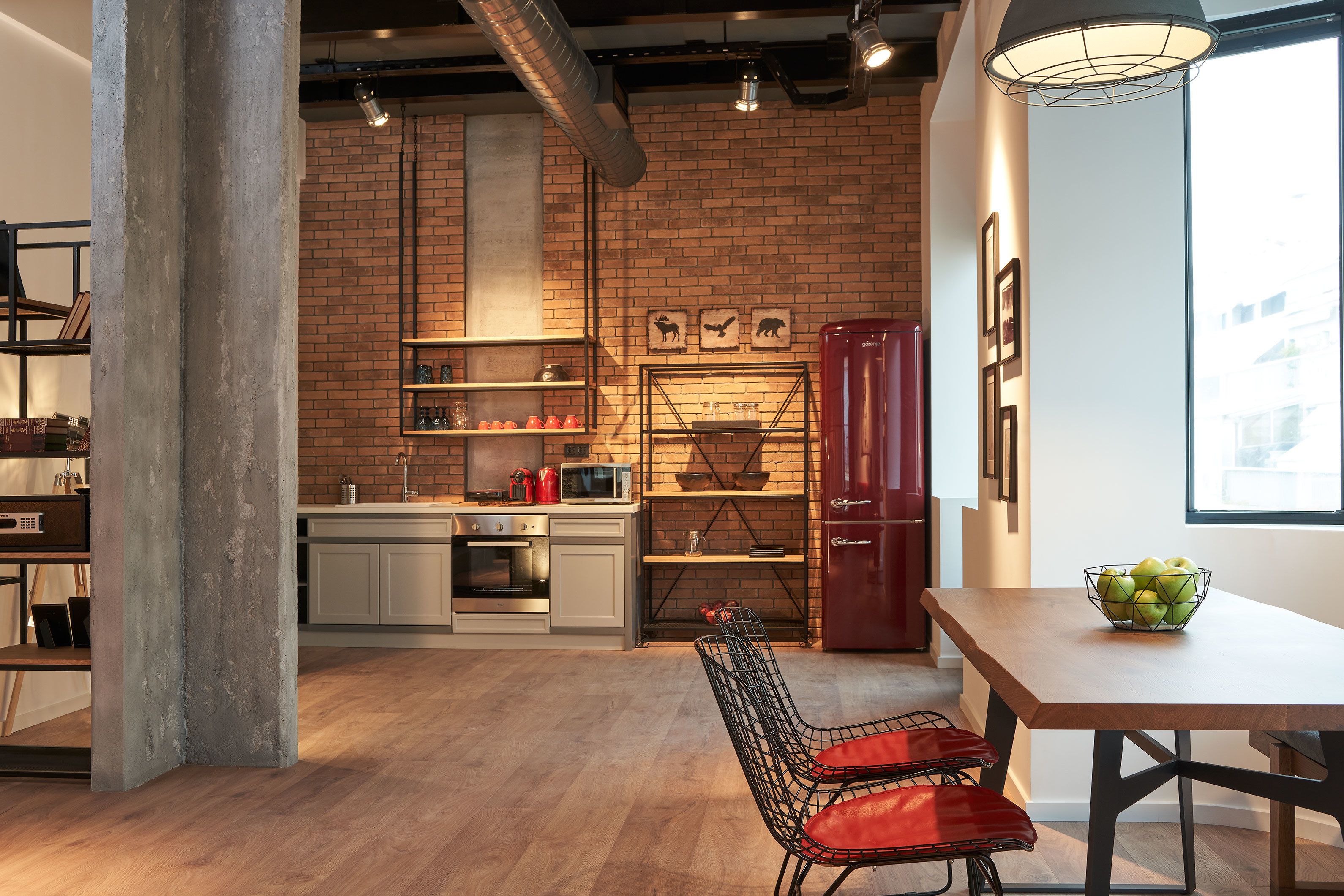 A modern kitchen with exposed brick walls, a wooden dining table, and a red refrigerator.