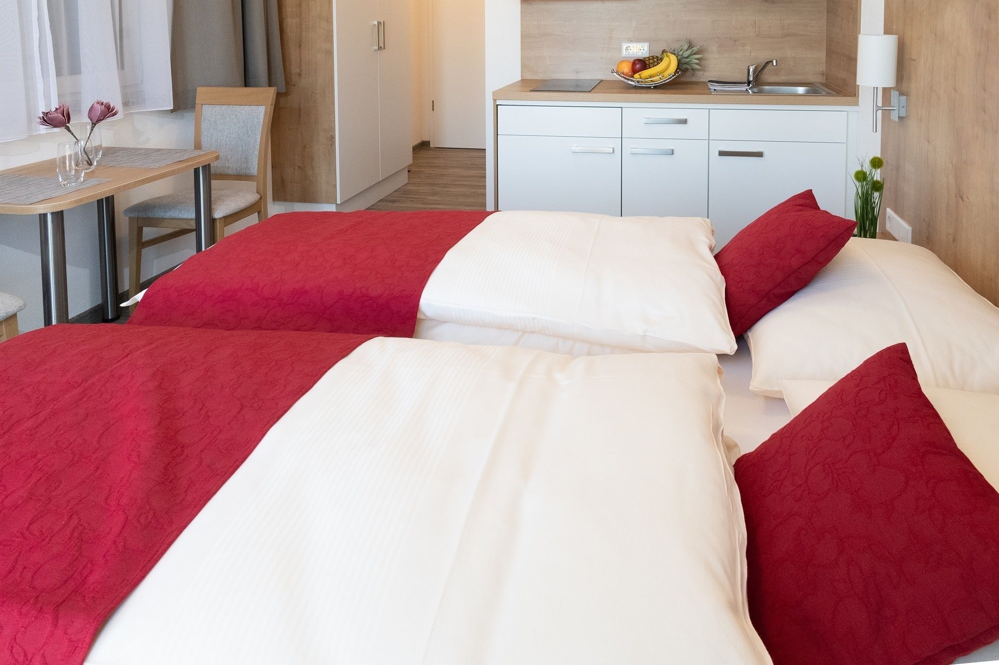A hotel room with two beds, a small table with a vase, a wooden chair, a kitchenette, and a window with curtains.