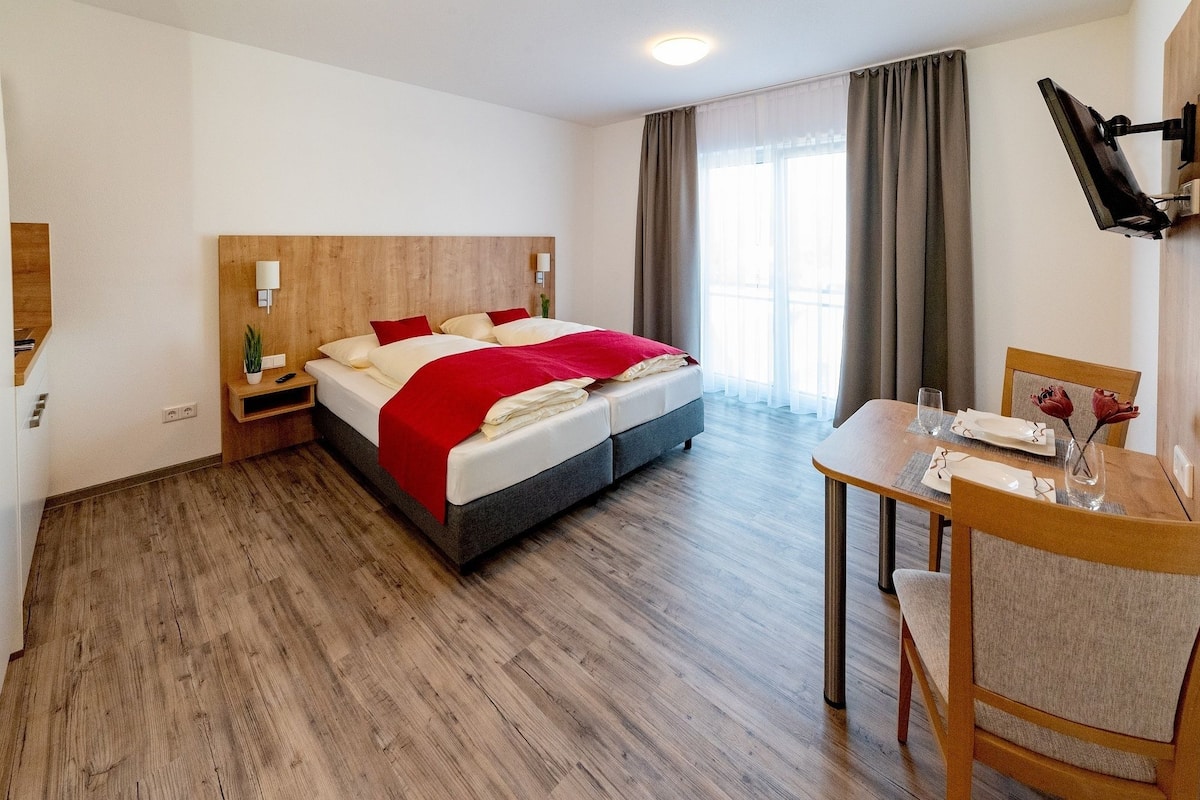 A hotel room with a bed, bedside table, lamp, and a dining area with a table and chairs.