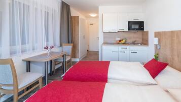 A hotel room with two beds, a dining table, a kitchen area, and a view of the hallway.