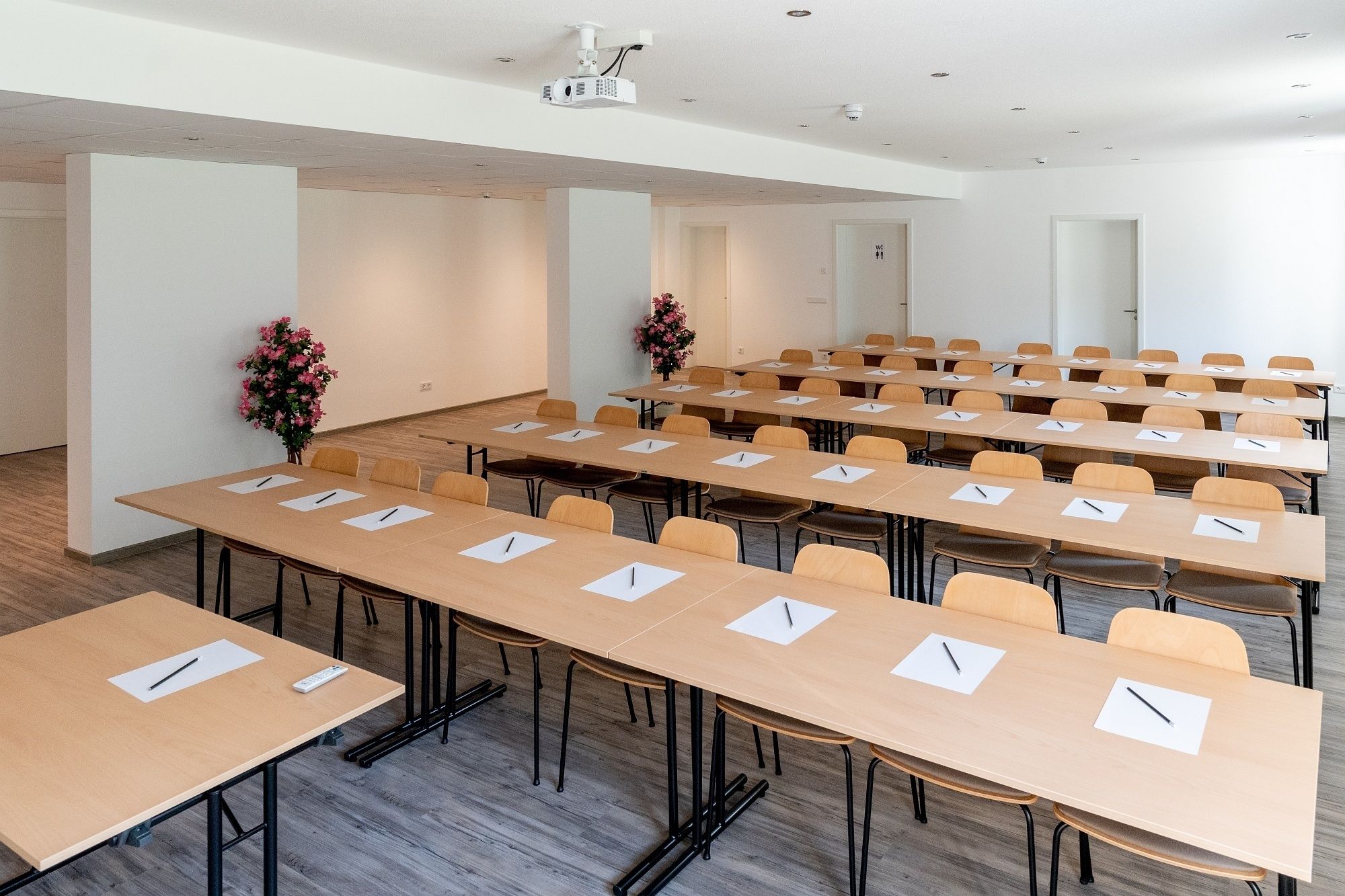 A conference room with multiple tables and chairs, equipped with pens and notepads, and a projector on the ceiling.