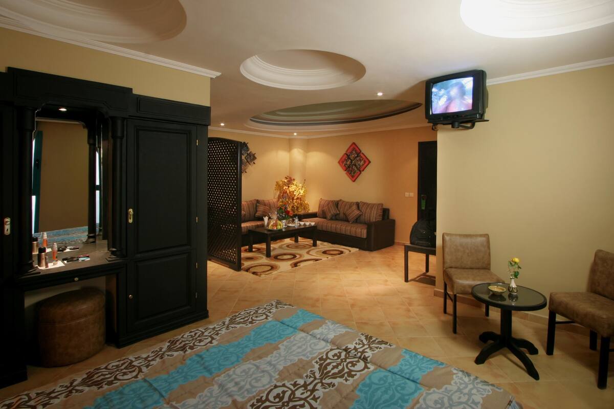 A hotel room with a large bed, a sitting area with a sofa and armchairs, a small table with a vase, and a television mounted on the wall.