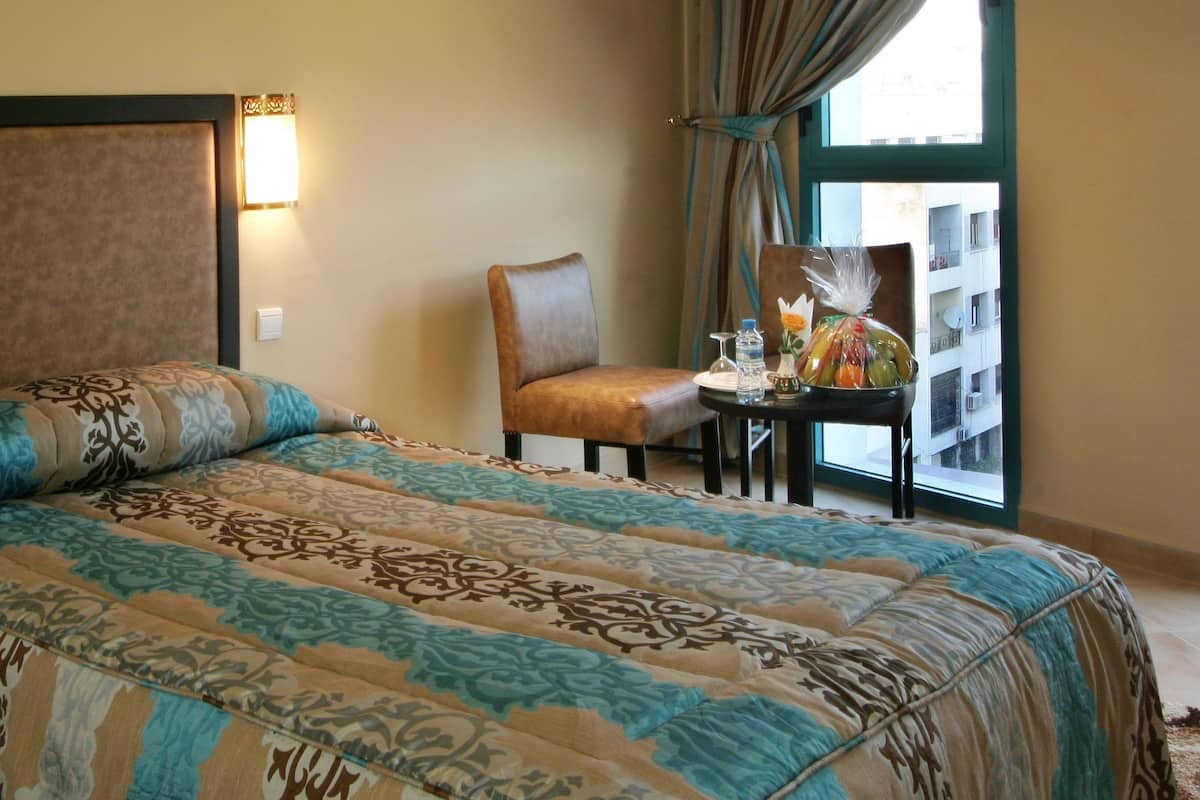 A hotel room with a large bed, a chair, a small table with a vase and snacks, and a window with curtains.