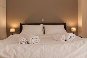 A neatly made bed with white linens and two rolled towels, a wooden headboard, and two bedside lamps.