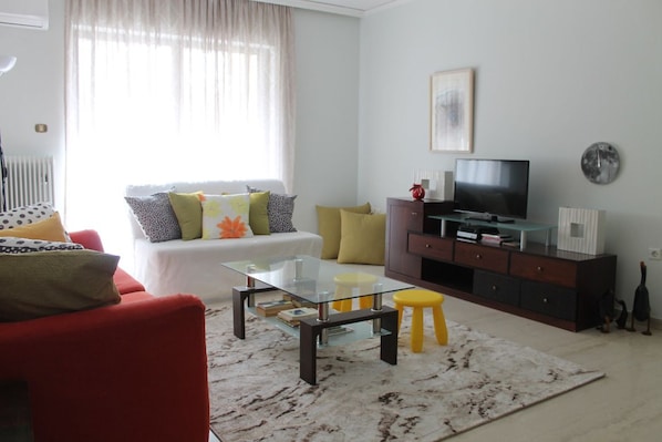 A living room with a glass coffee table, red sofa, and yellow stool. There is a TV on a wooden cabinet and a window with curtains.