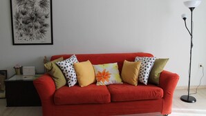 Ein rotes Sofa mit vielen Kissen, ein gerahmtes Kunstwerk an der Wand, eine Stehlampe und ein kleiner Tisch mit einer Blumenvase.