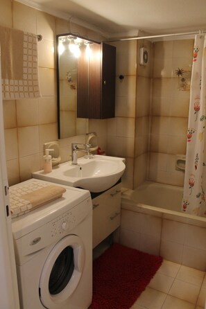 A compact bathroom with a washing machine, sink, mirror, and shower.