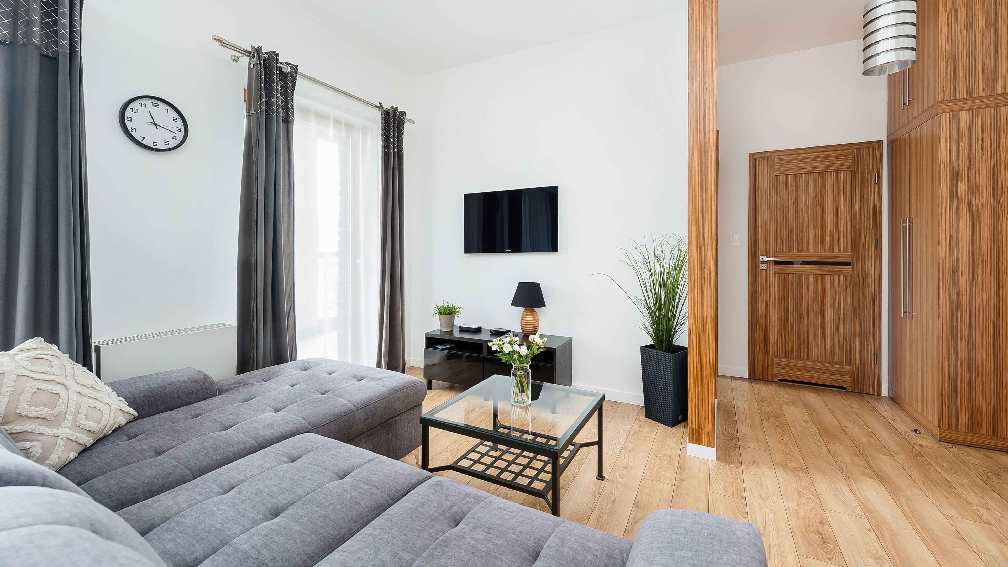 A modern living room with a grey sofa, a glass coffee table, a TV on the wall, and a wooden wardrobe.