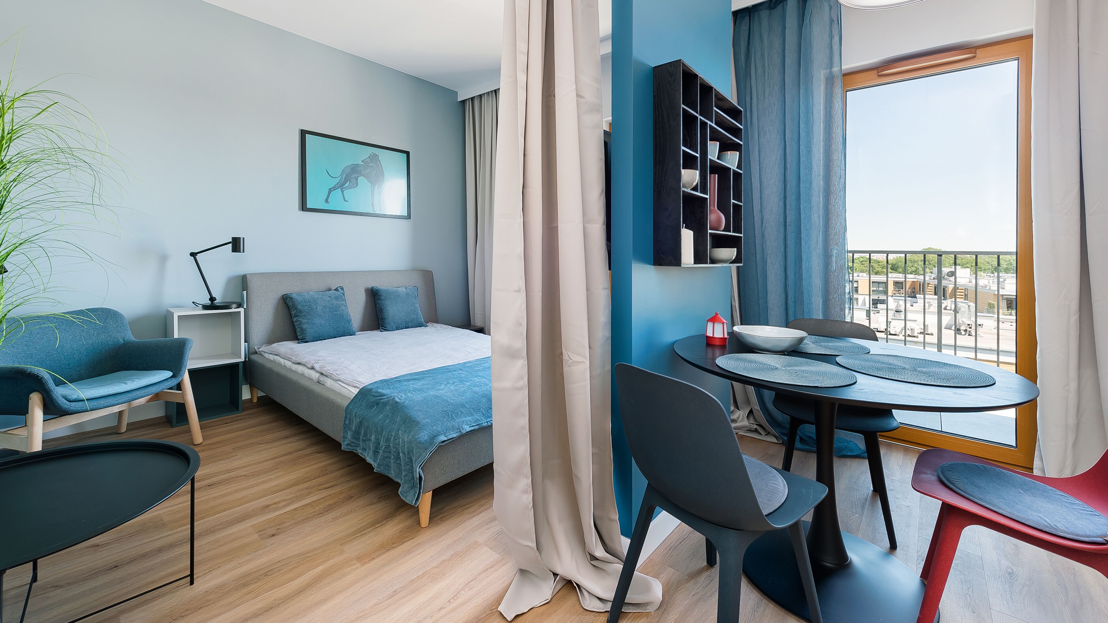 A modern bedroom with a bed, a desk, chairs, a dining table, and a view of the outside.