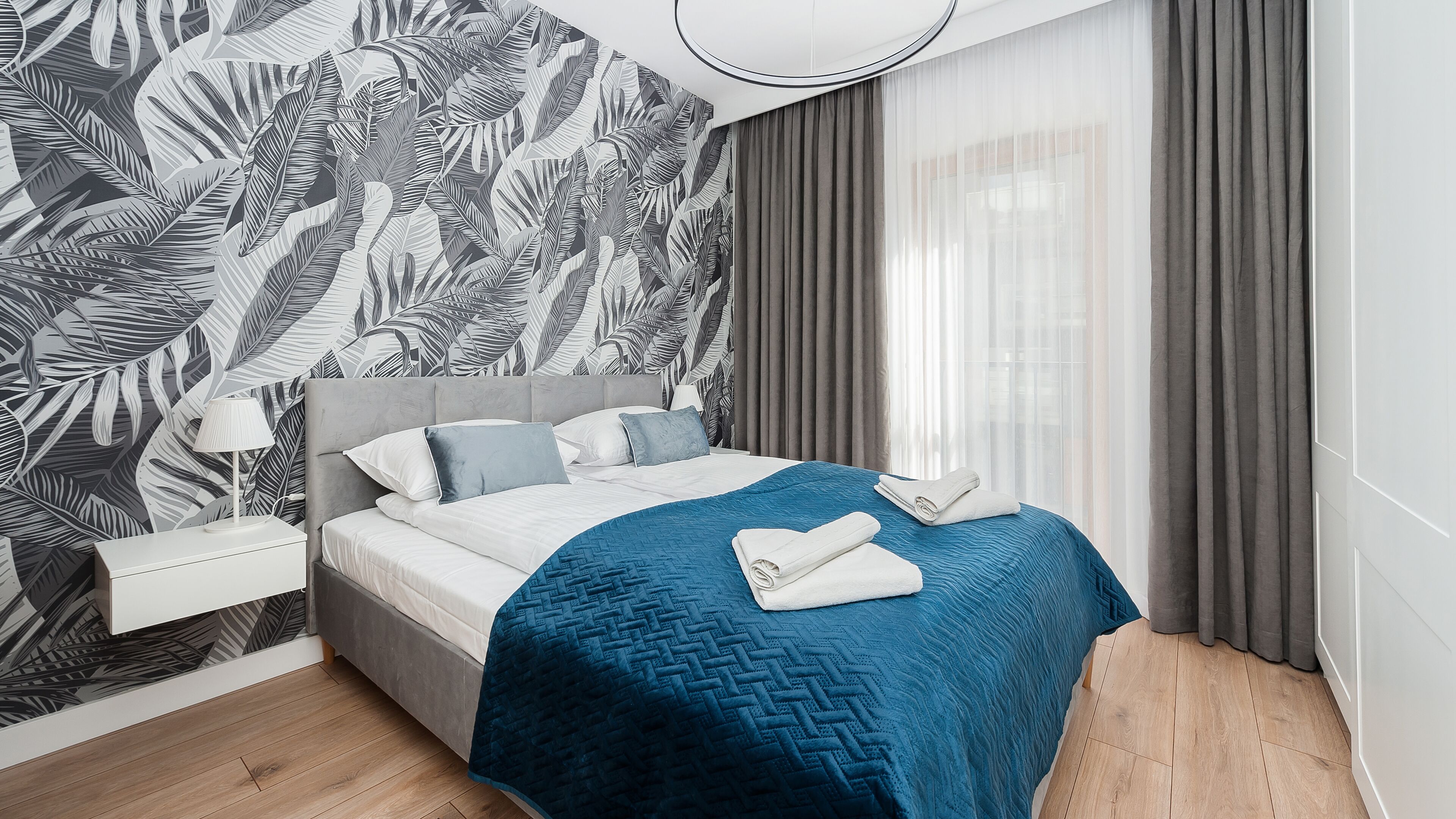 A modern hotel room with a large bed, a nightstand, a lamp, and a tropical leaf-patterned wall.