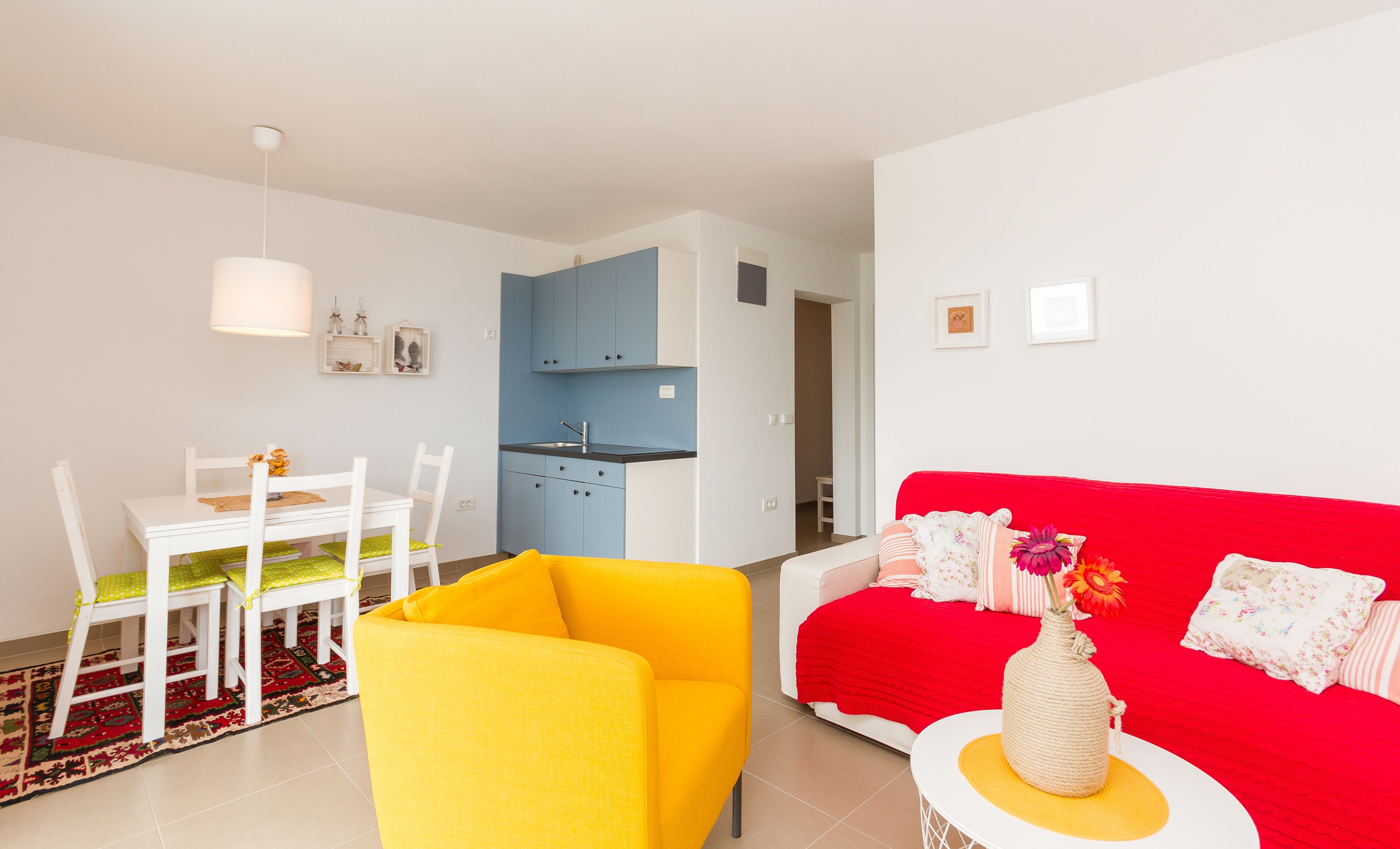 A bright living room with a yellow armchair, a red sofa, and a white table with chairs. There is a kitchen area with blue cabinets and a white ceiling light.