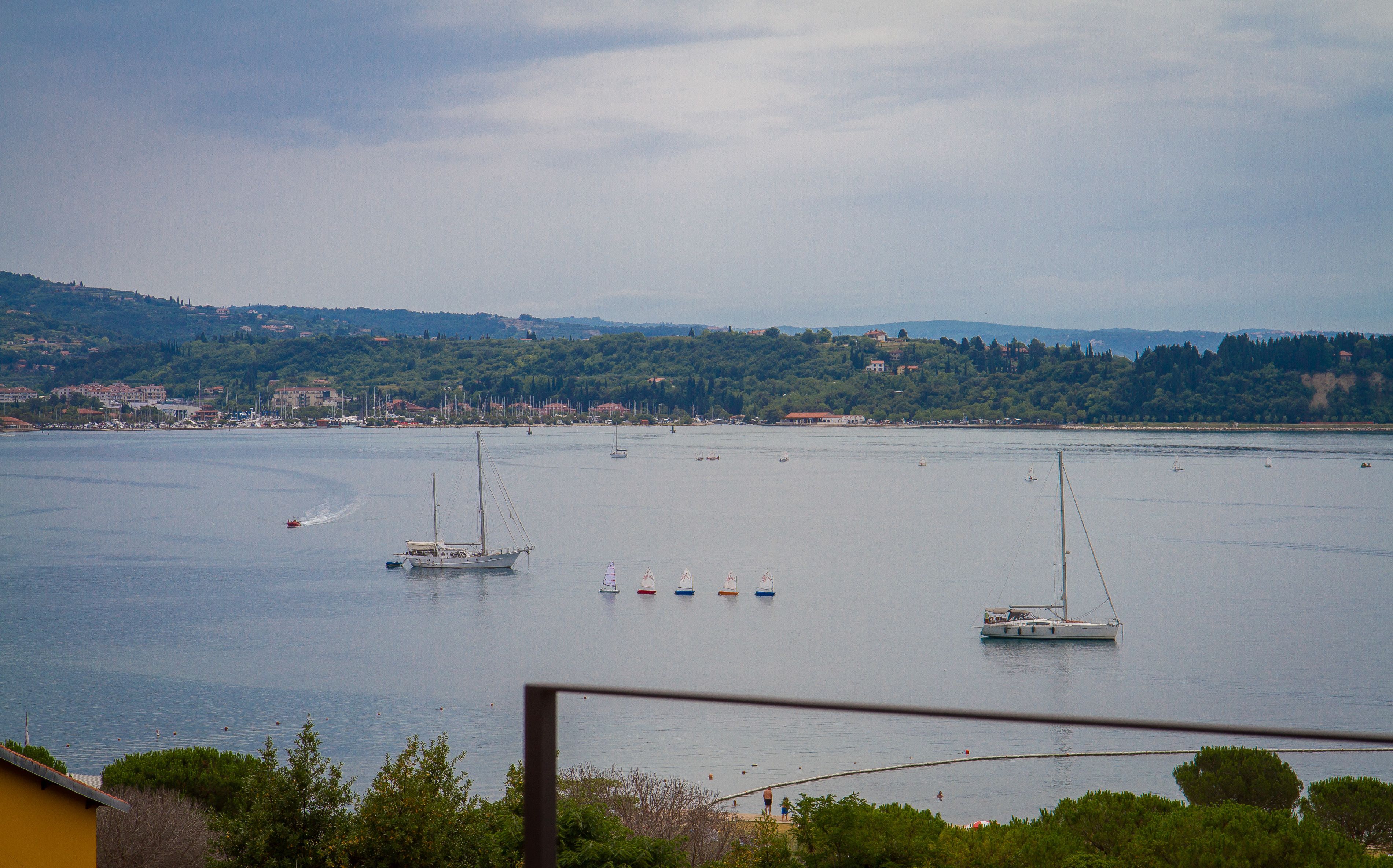 A coastal view with boats on the water, a distant shoreline with buildings, and a hilly landscape.
