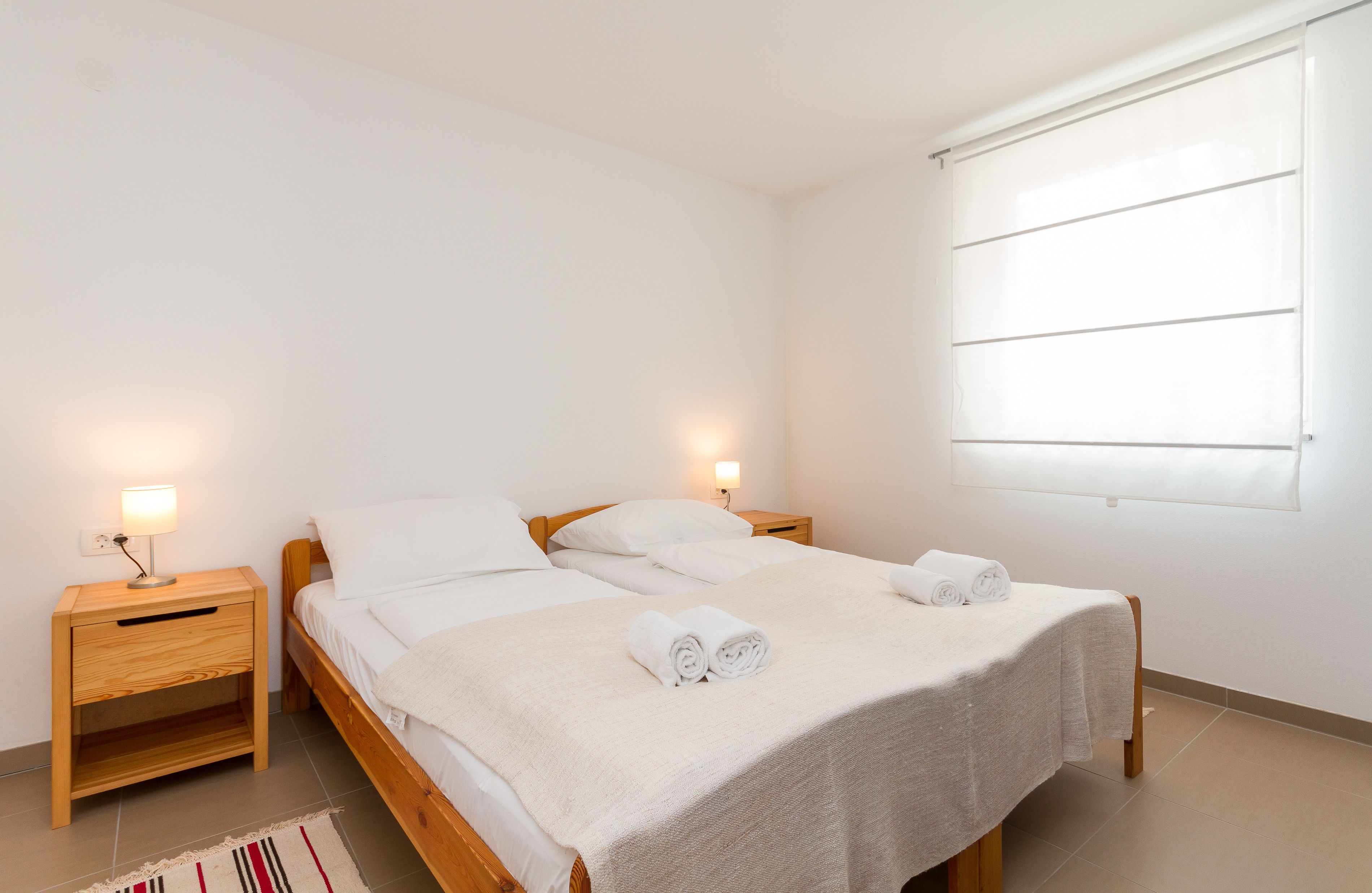 Una cama doble con dos almohadas, una mesita de noche con lámpara y una ventana con persianas.