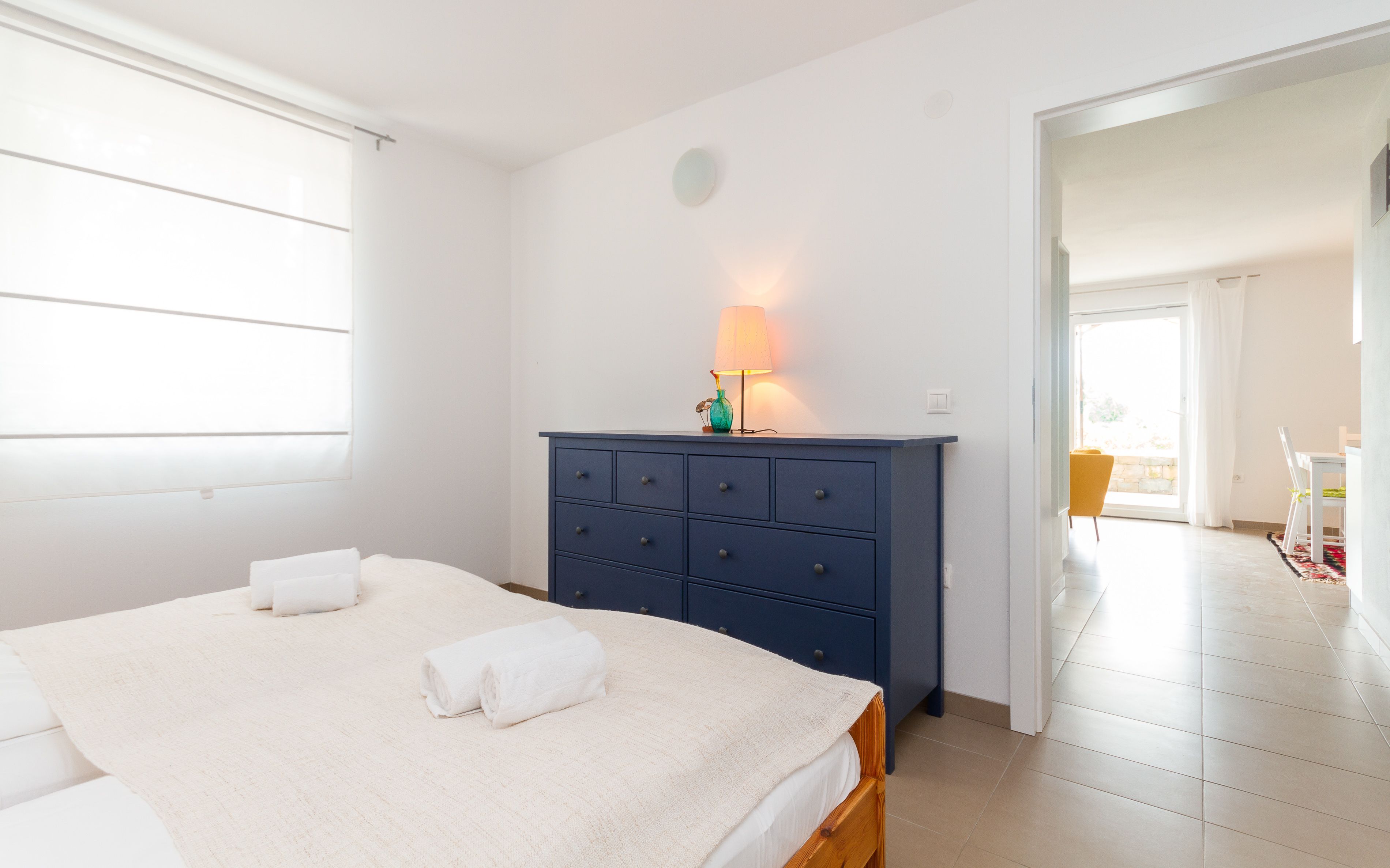 Un dormitorio con cama, cómoda, lámpara y vista al exterior.