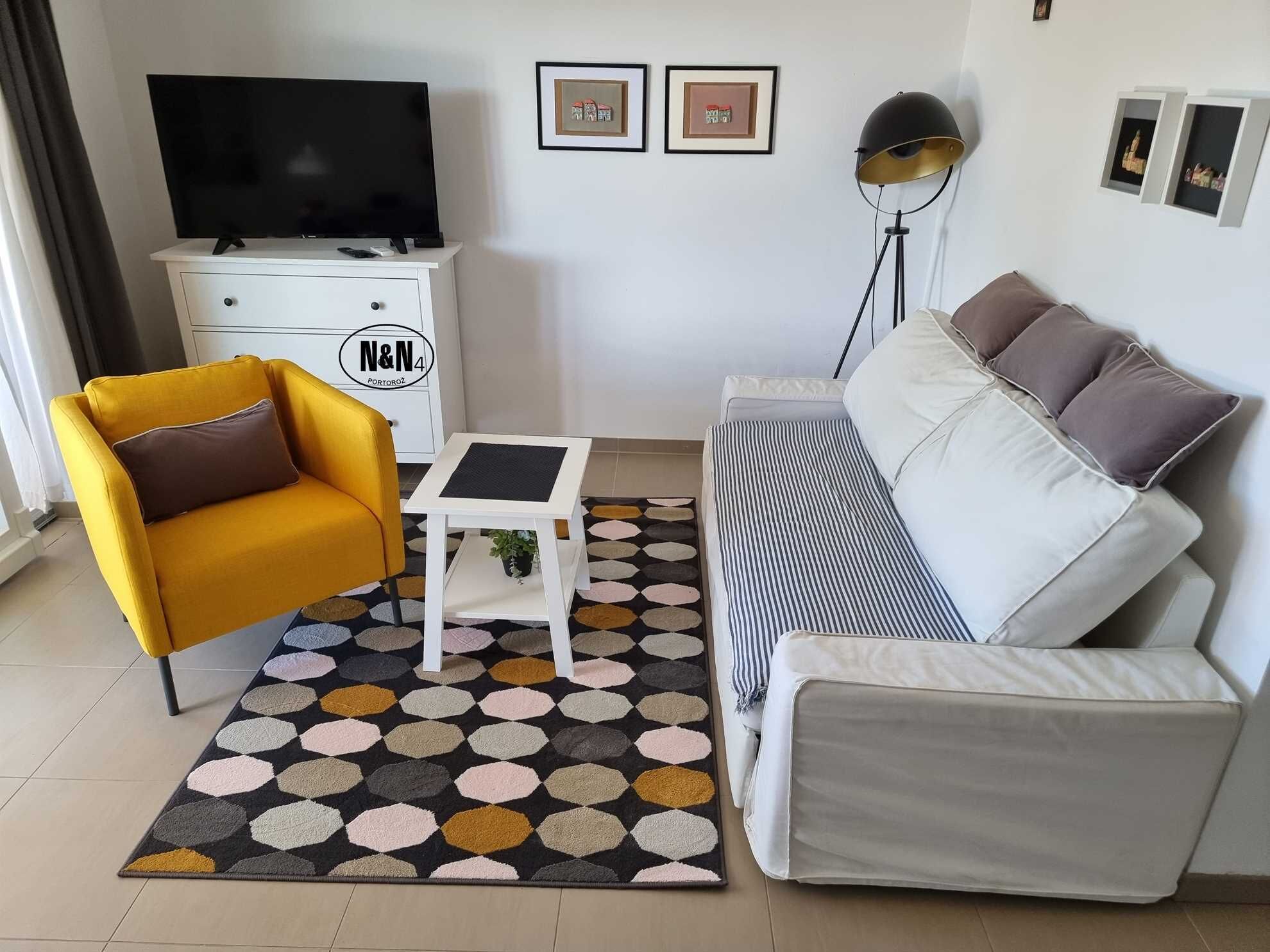 A living room with a yellow armchair, a white sofa, a small white table, a black TV on a white cabinet, and a patterned rug.