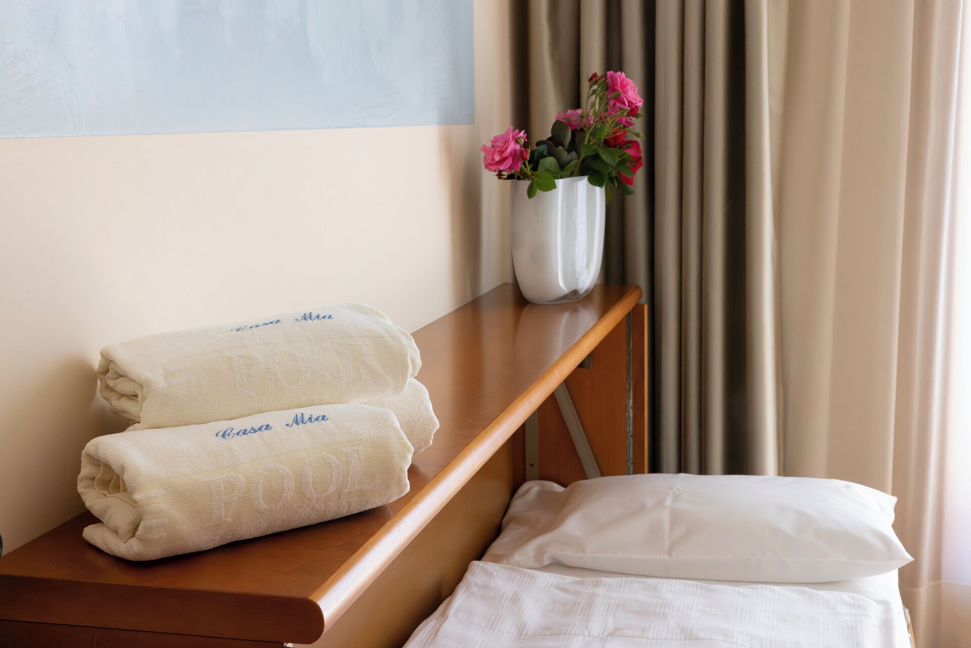 Ein ordentlich gemachtes Bett mit weißen Leinen, ein Nachttisch aus Holz mit gefalteten Handtüchern und eine Vase mit rosa Blumen darauf.