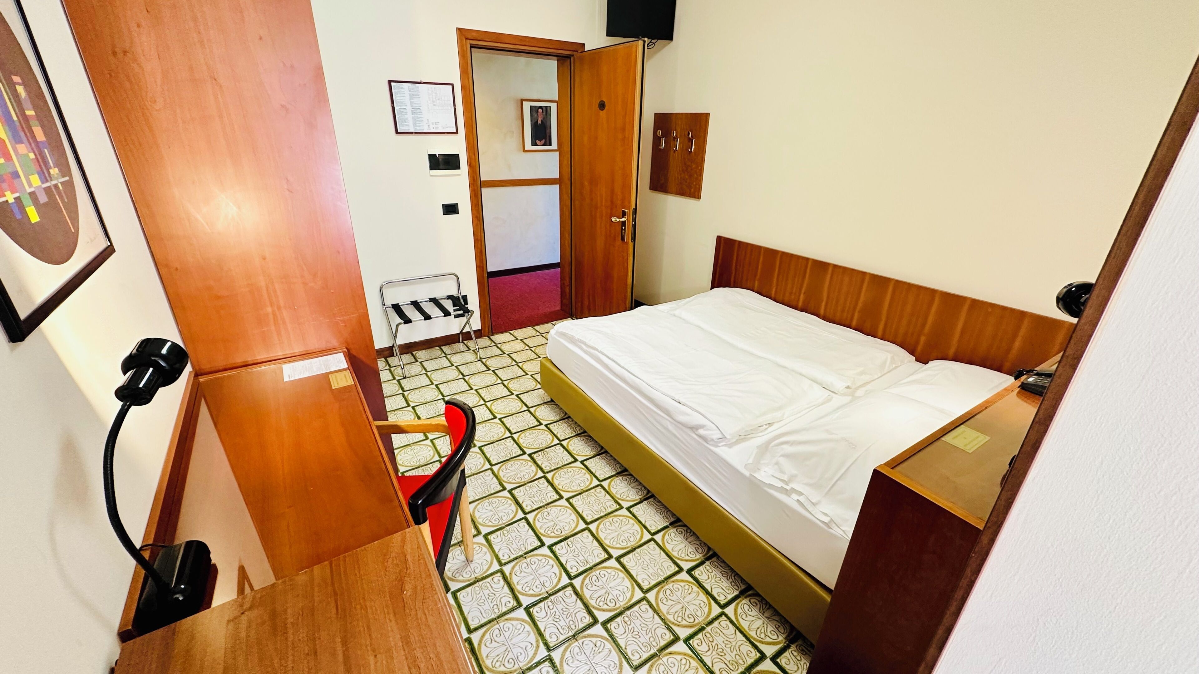 A room with a bed, a desk, a chair, a wardrobe, and a door leading to another room.