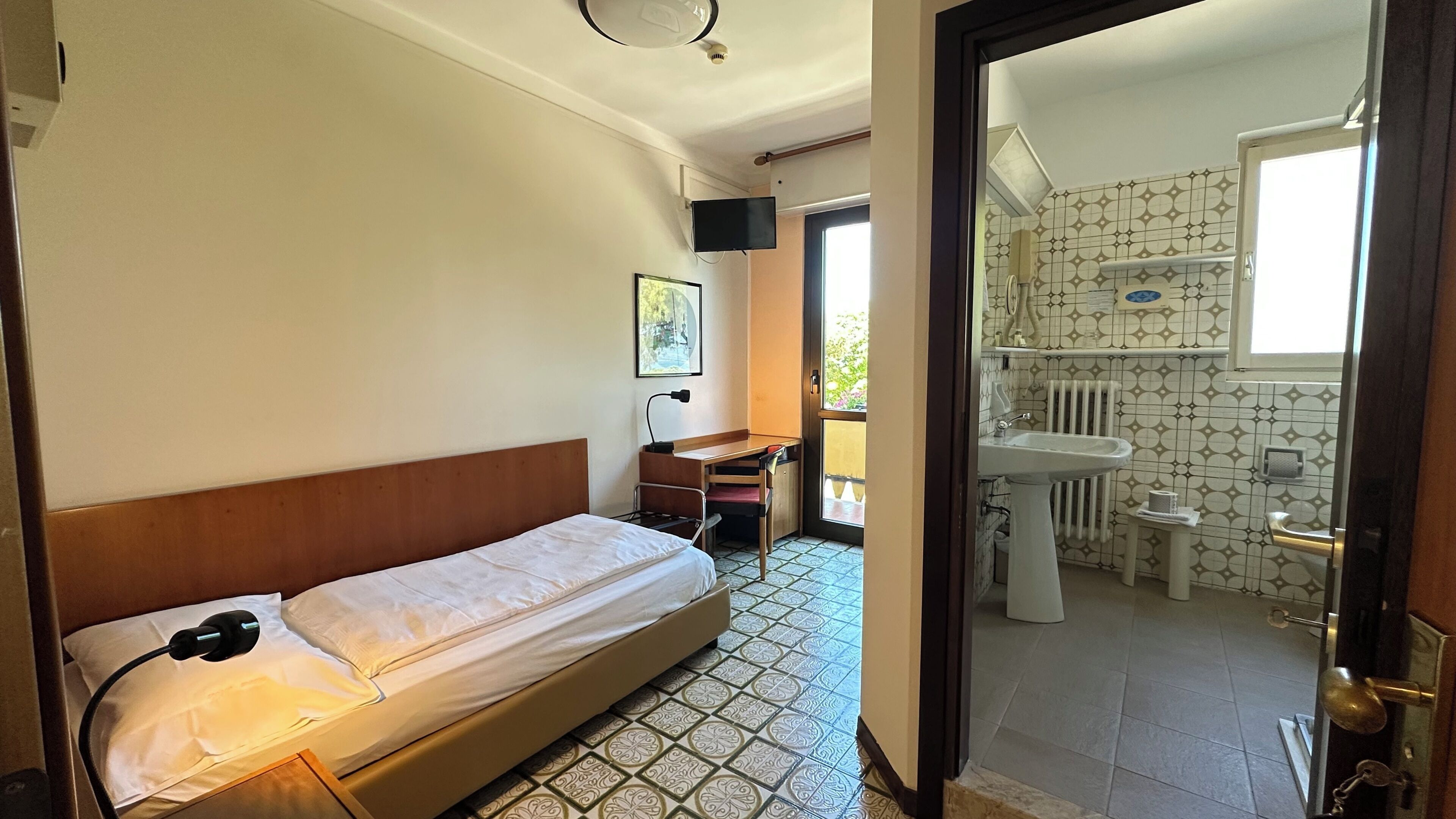 A bedroom with a bed, a desk, a chair, a TV, and a bathroom with a sink and toilet.